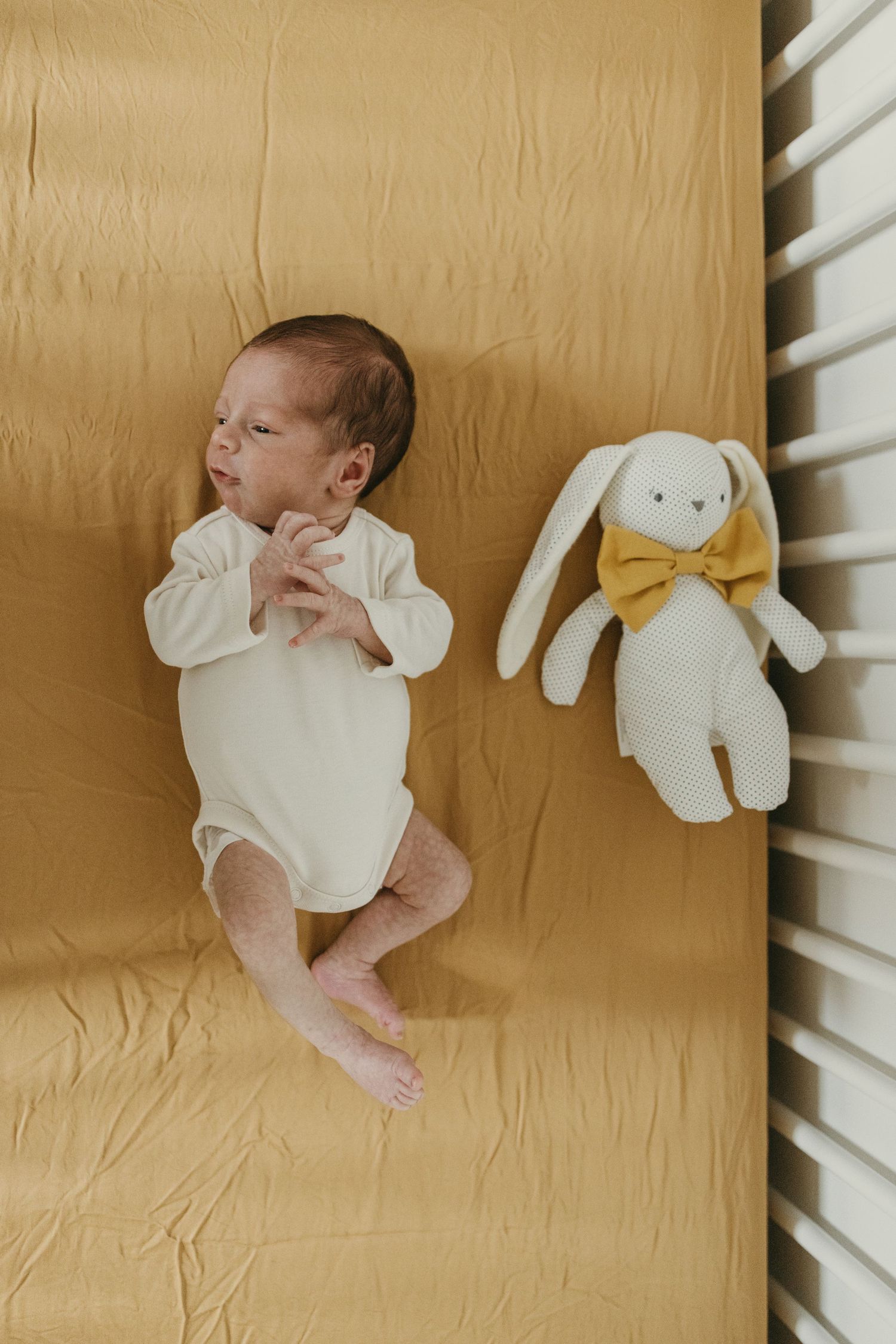 Baby in white onesie lying on golden blanket next to plush bunny with yellow bow.