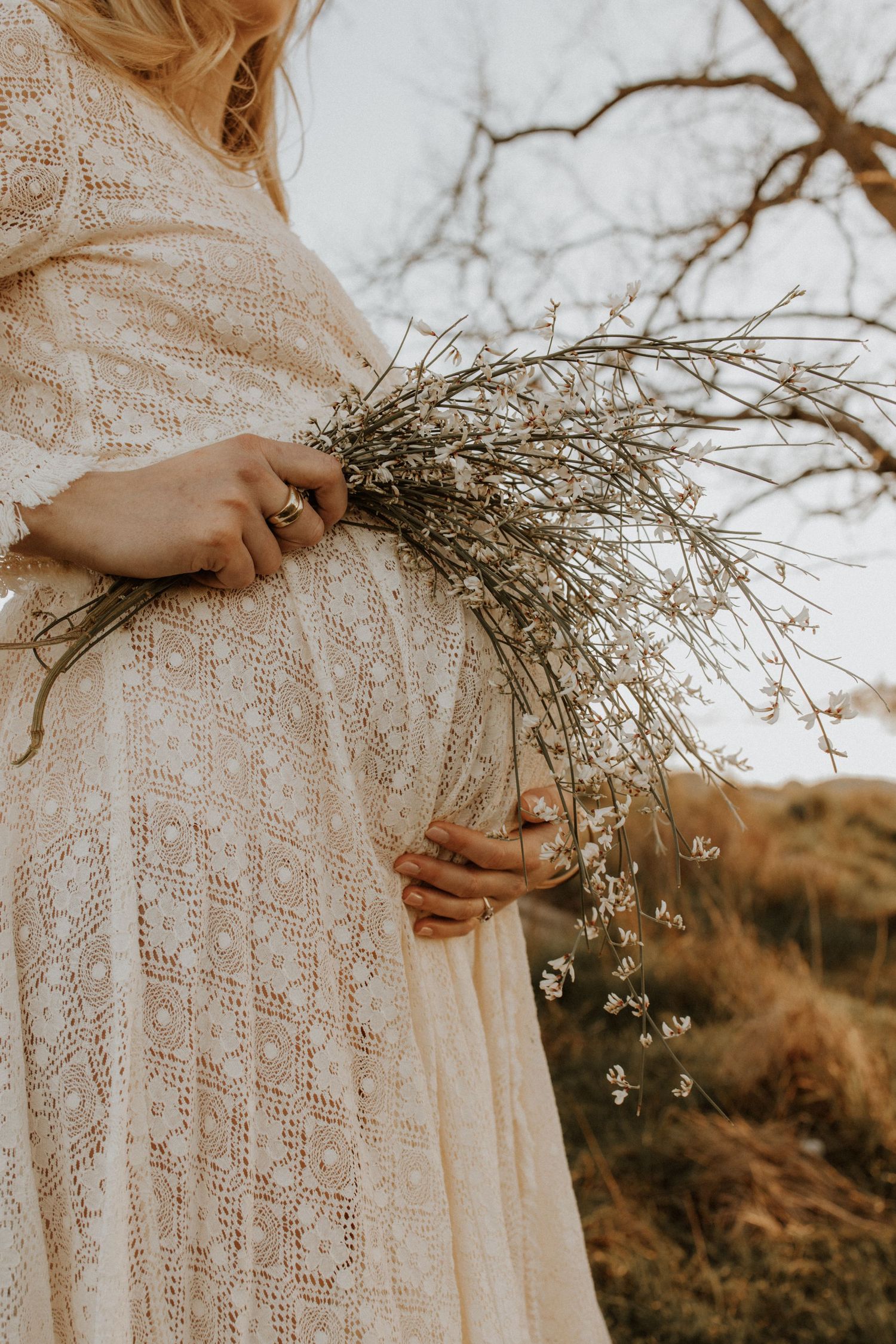 A person in a lace dress holds dried flowers while standing outdoors in golden light.
