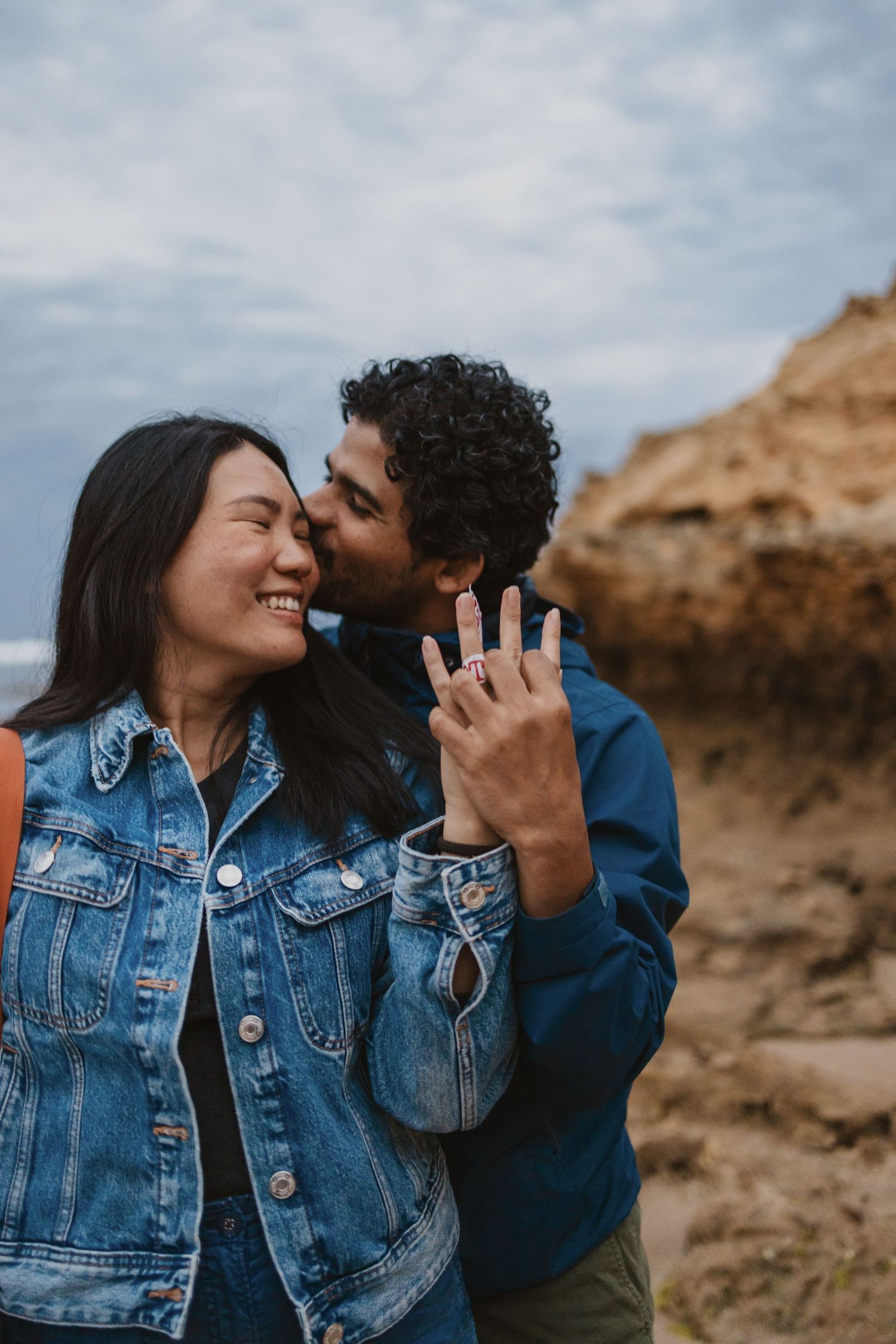 Couple celebrating engagement outdoors, woman showing ring while man kisses her cheek, both wearing denim jackets near rocky cliff.