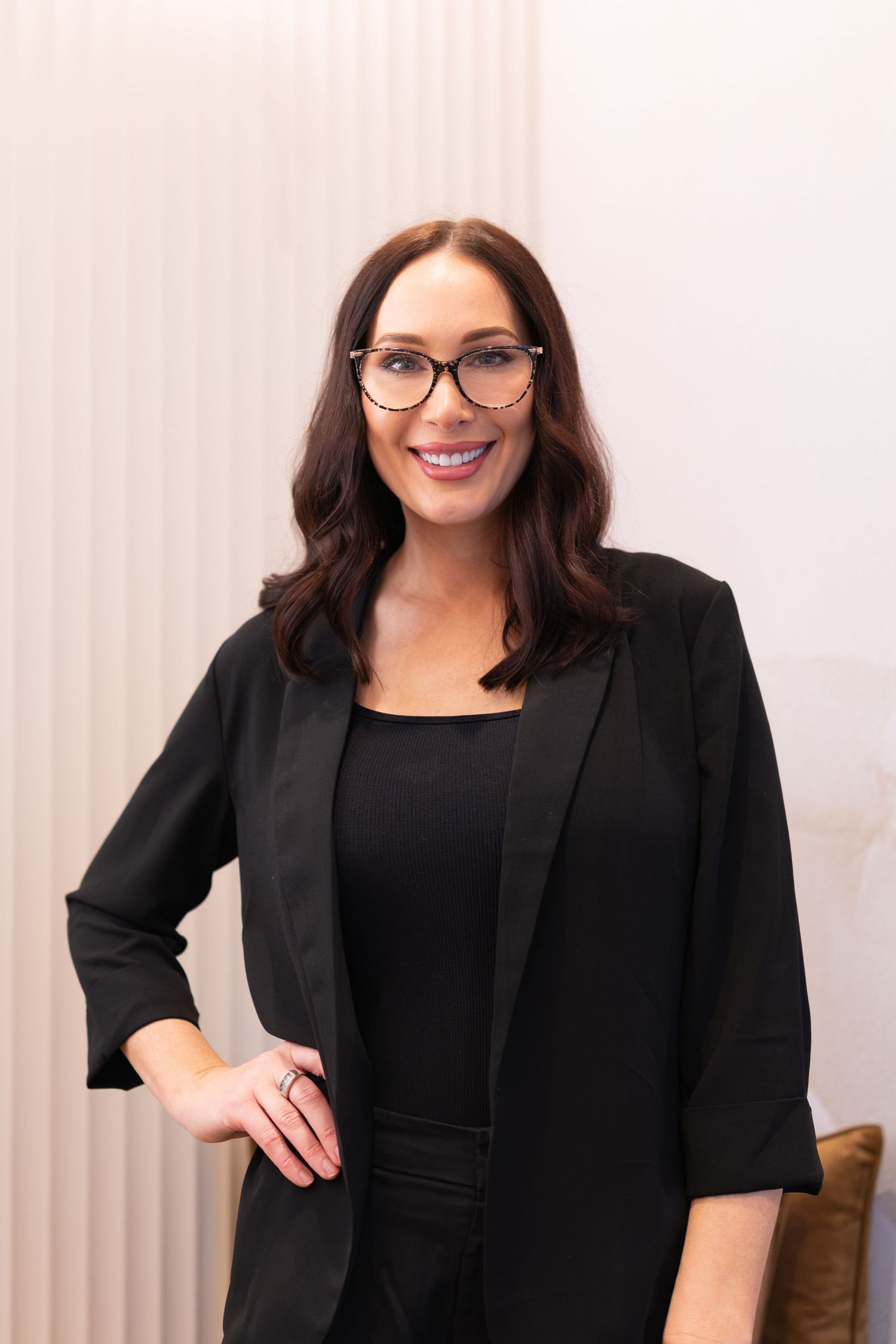 Professional woman in black blazer and glasses smiling confidently in a modern office setting.