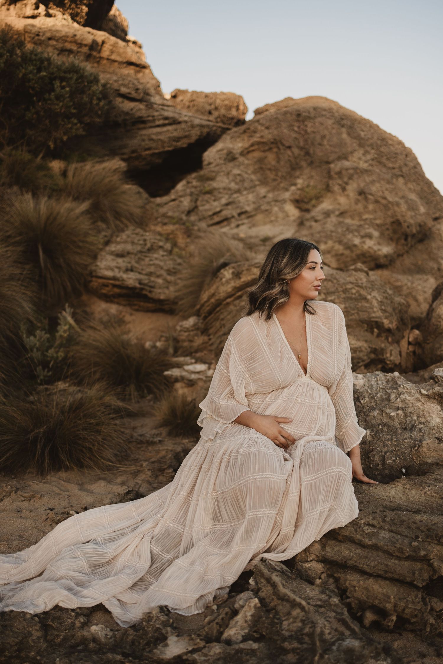 Pregnant woman in flowing cream boho dress sitting on desert rocks, smiling serenely at golden hour.
