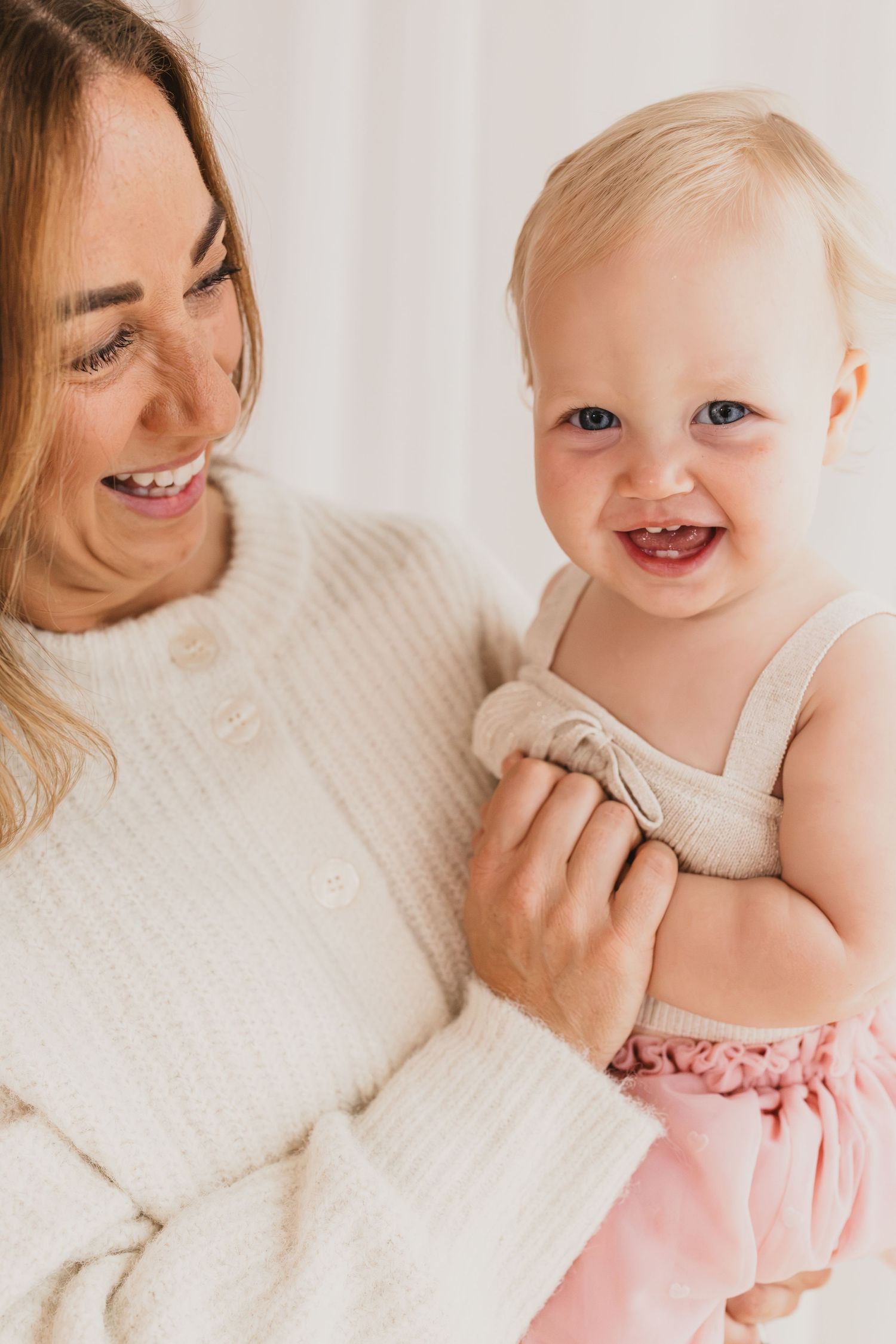 Smiling mother in white sweater holding her happy blue-eyed baby girl in pink dress.