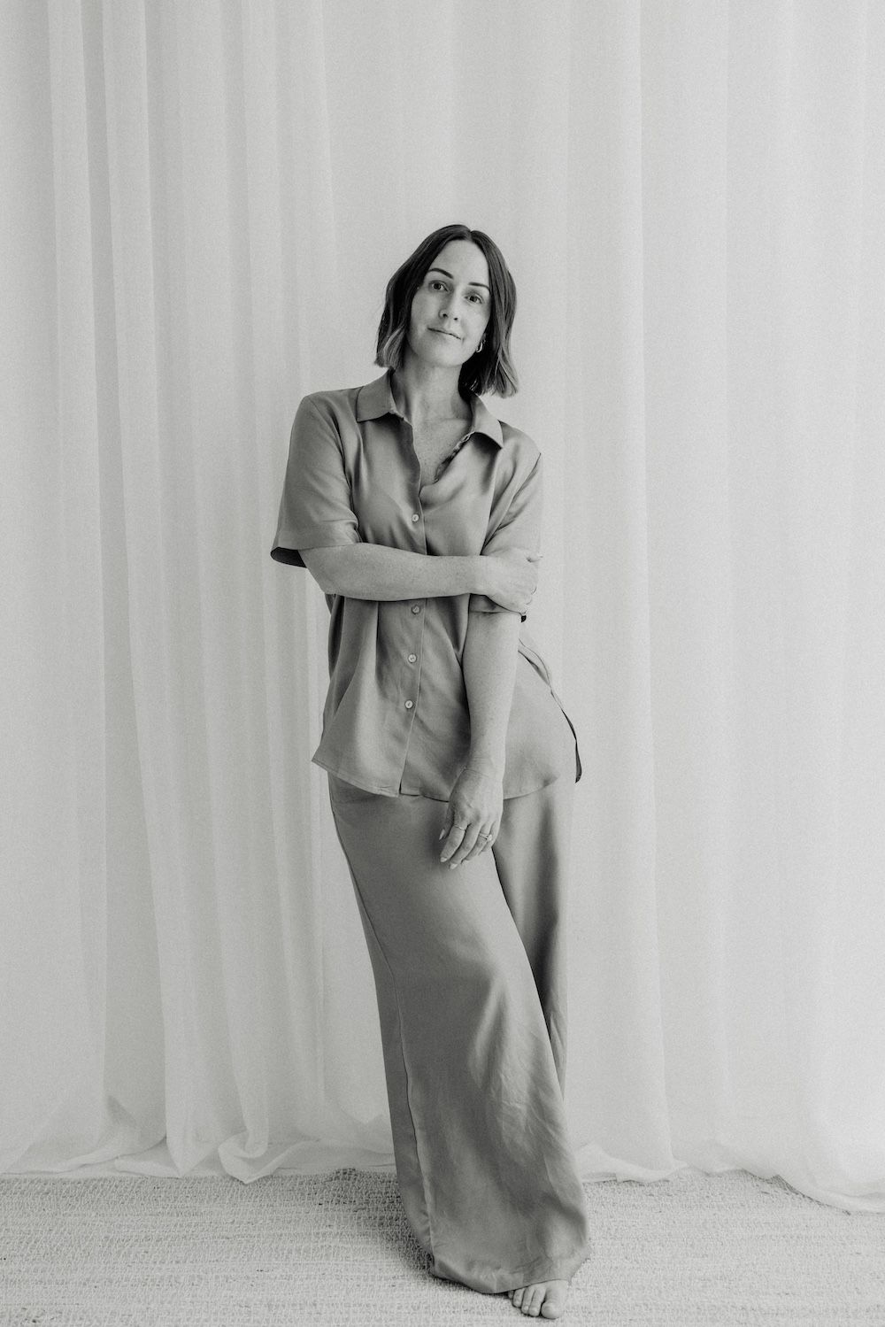 Woman in satin pajama set poses gracefully against sheer white curtains in a black and white portrait photo.