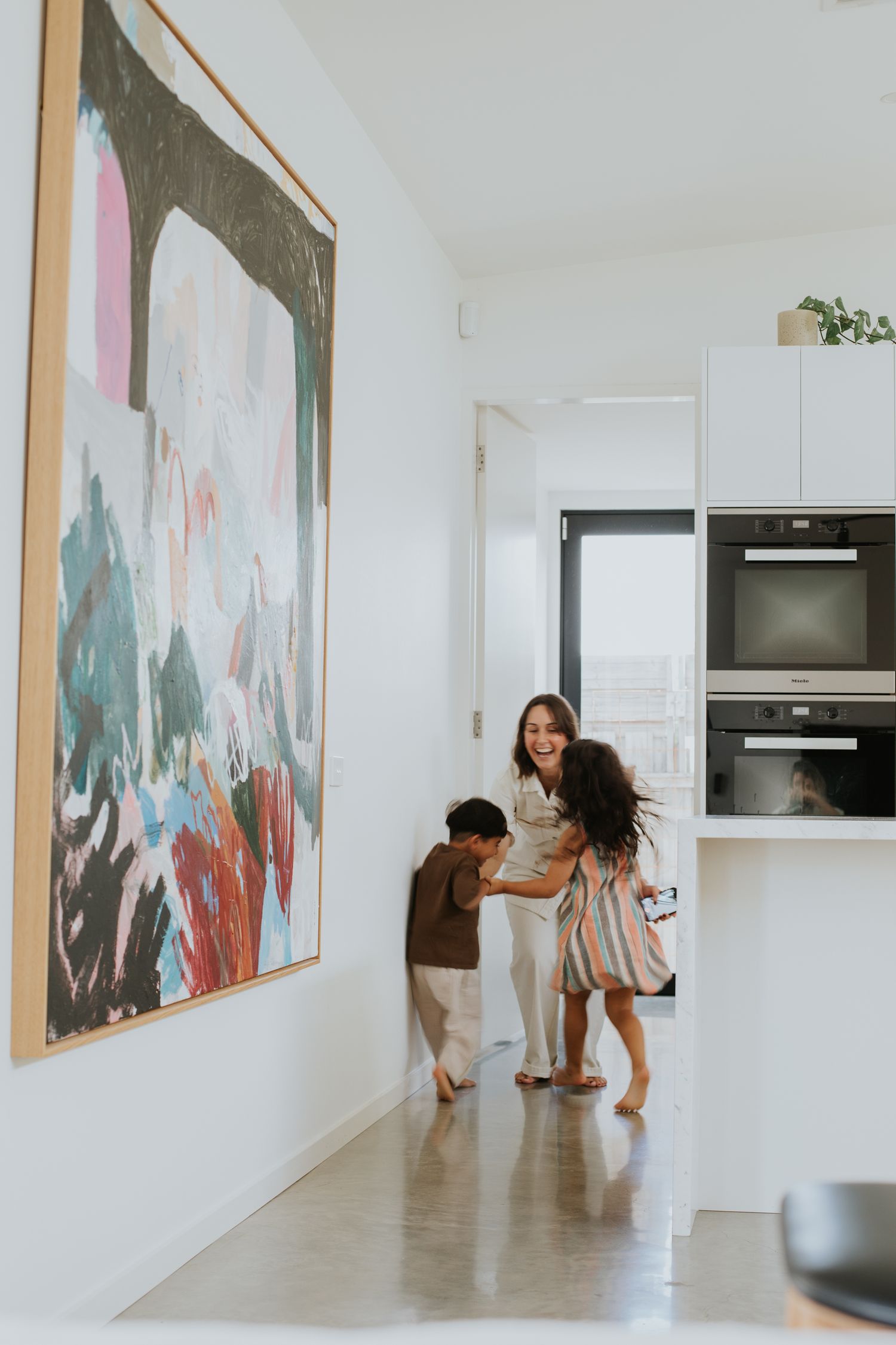 A mother plays joyfully with her two children in a modern white kitchen beside a large colorful abstract painting.