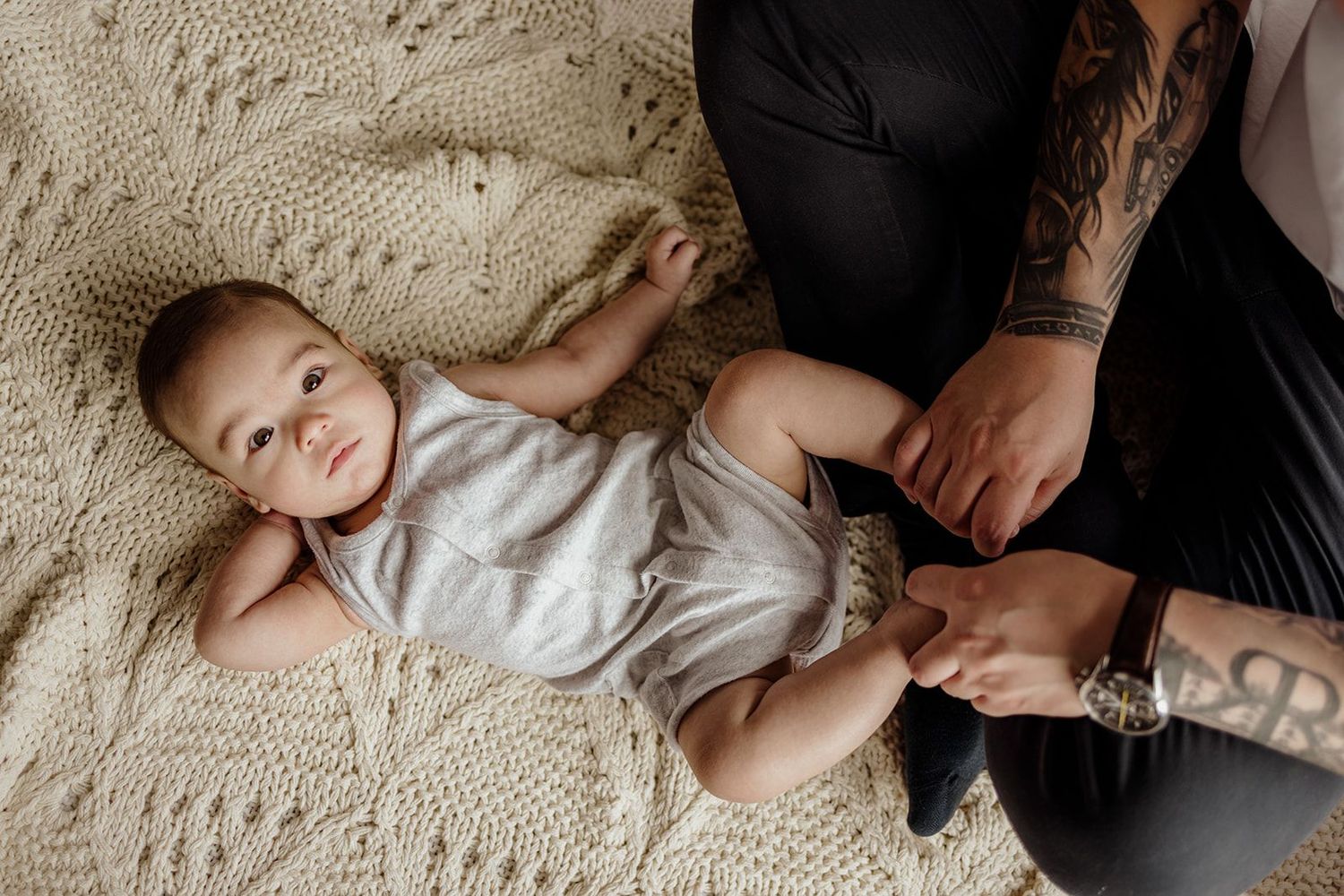 Tattooed parent changing baby's diaper on a knit blanket, infant in white onesie looking up curiously.