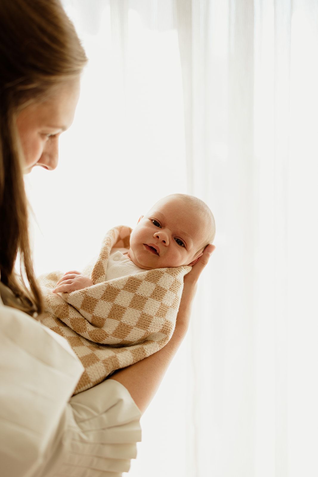 A mother tenderly holds her newborn baby wrapped in a beige checkered blanket near a bright window.