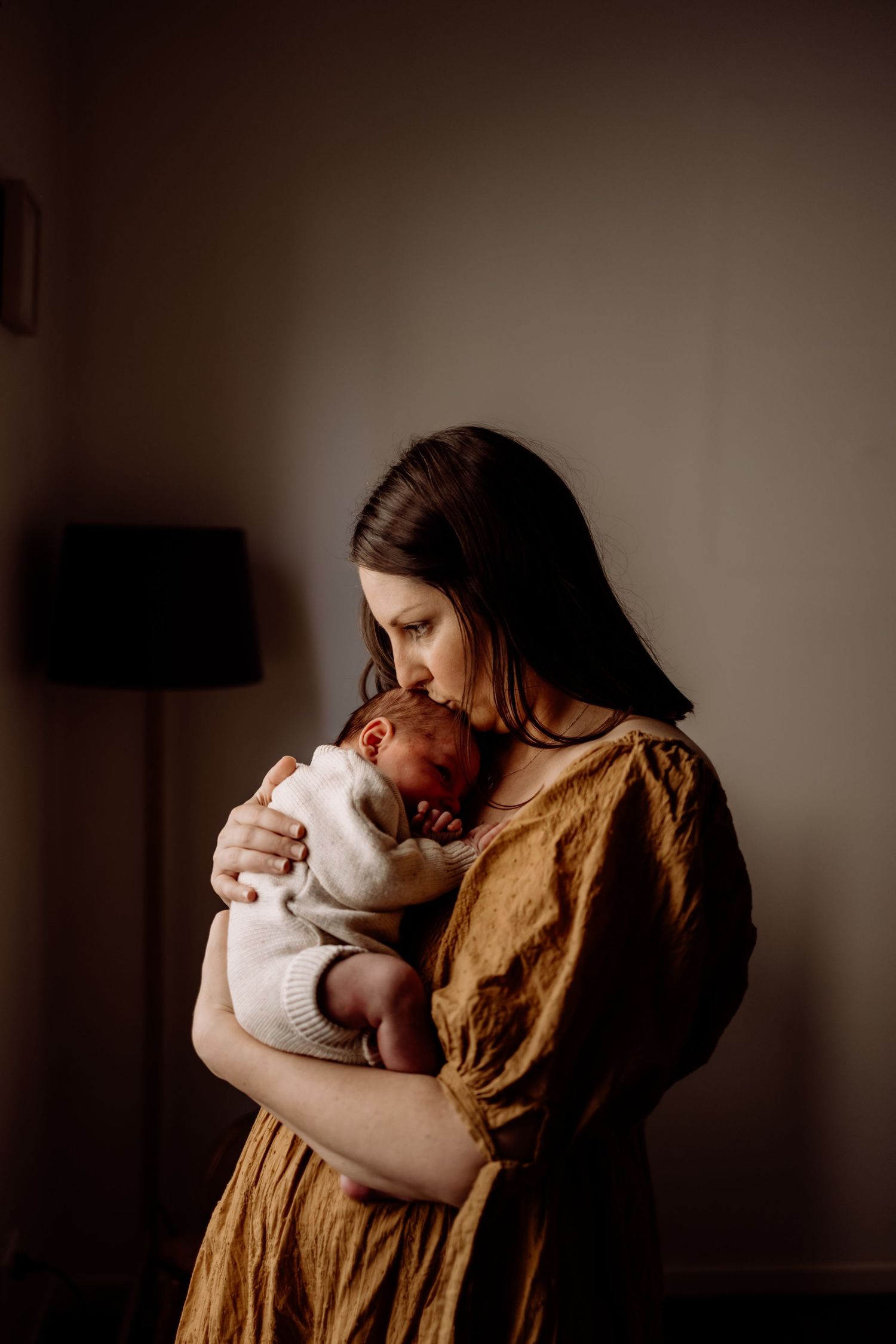 A mother in a mustard dress tenderly holds and kisses her sleeping newborn baby in soft, warm indoor lighting.