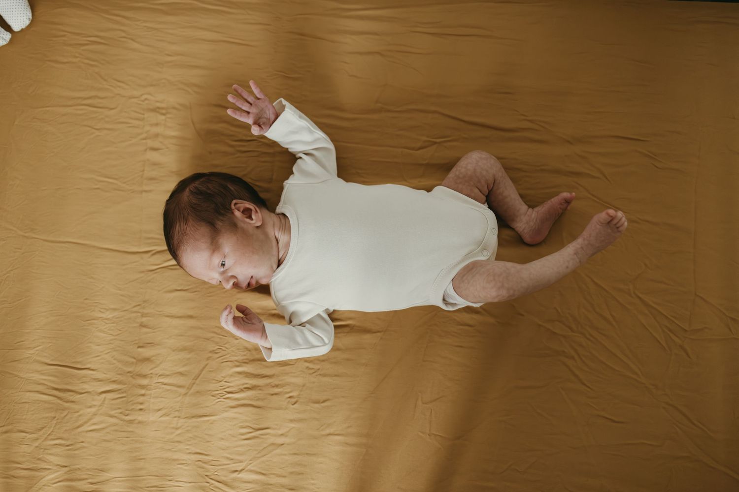 Baby in white onesie lying on golden brown blanket with arms stretched out.