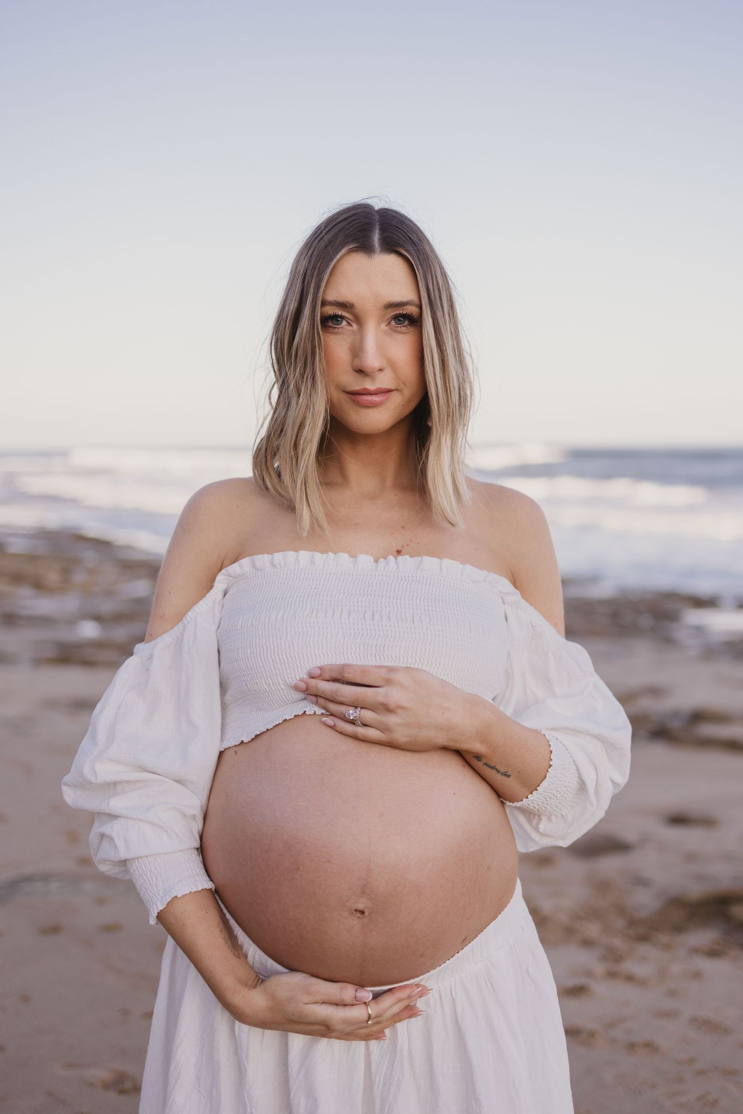 Pregnant woman in white off-shoulder dress cradling bare baby bump on sandy beach with ocean waves behind her.