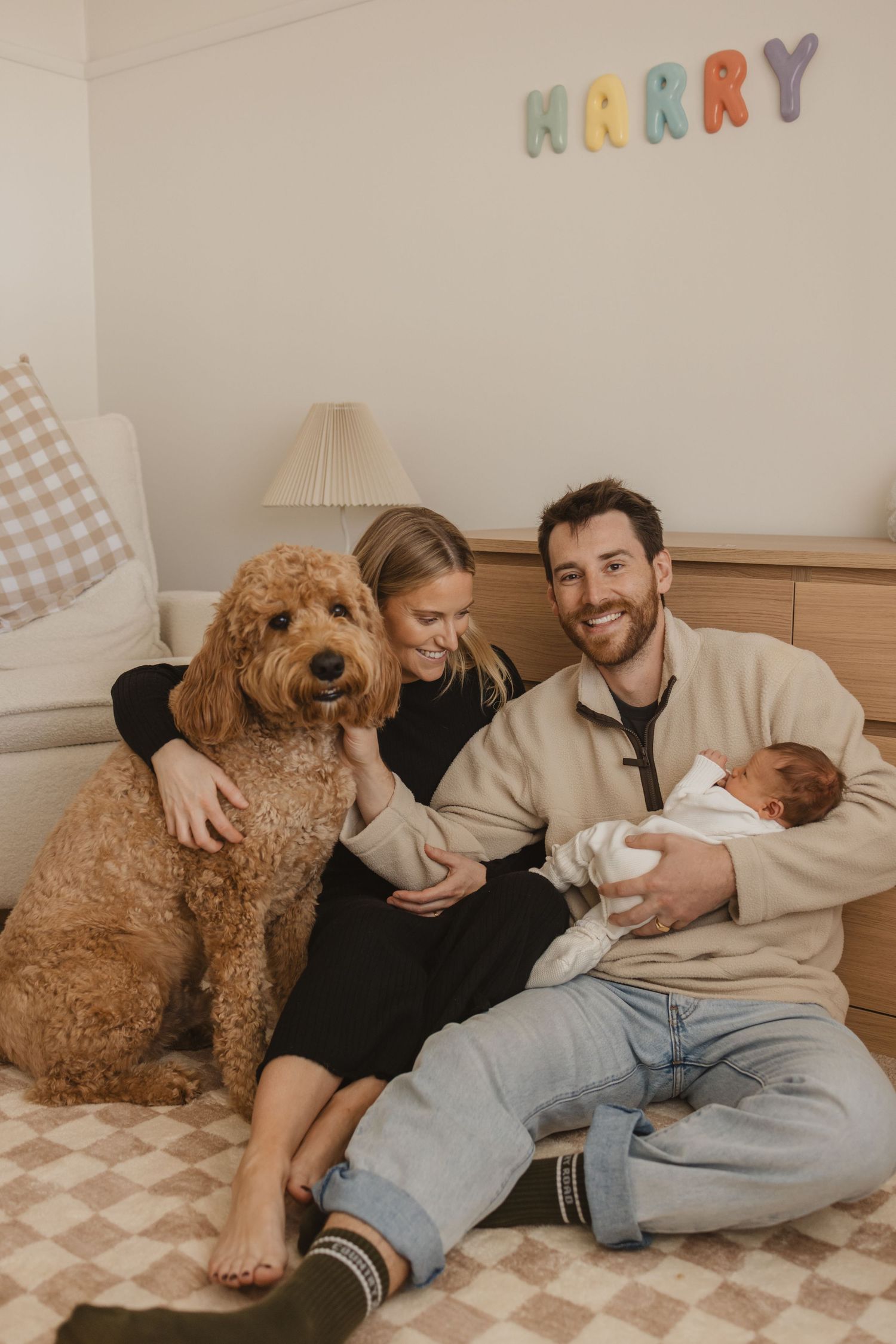 Happy couple sitting on floor with golden doodle dog and newborn baby under colourful HARRY letters.