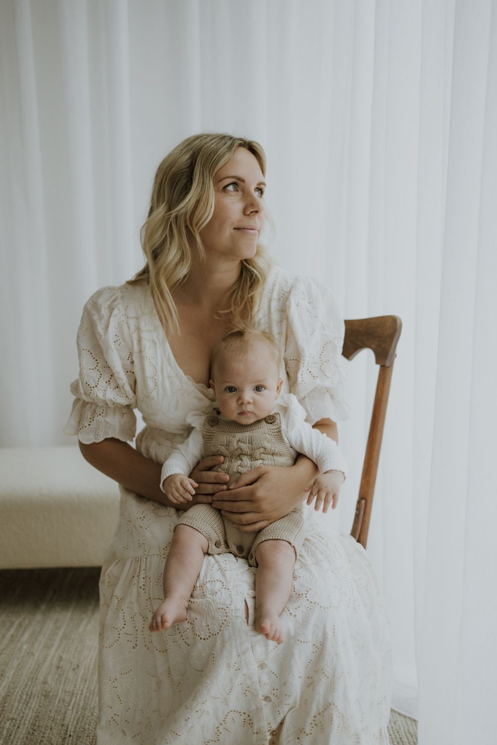 Mother and baby in white lace dress sitting on wooden chair by window.