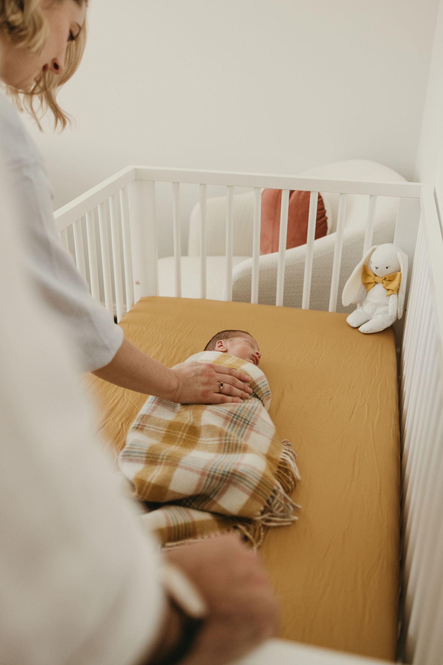 Baby sleeping in white crib with plaid blanket and stuffed bunny toy.
