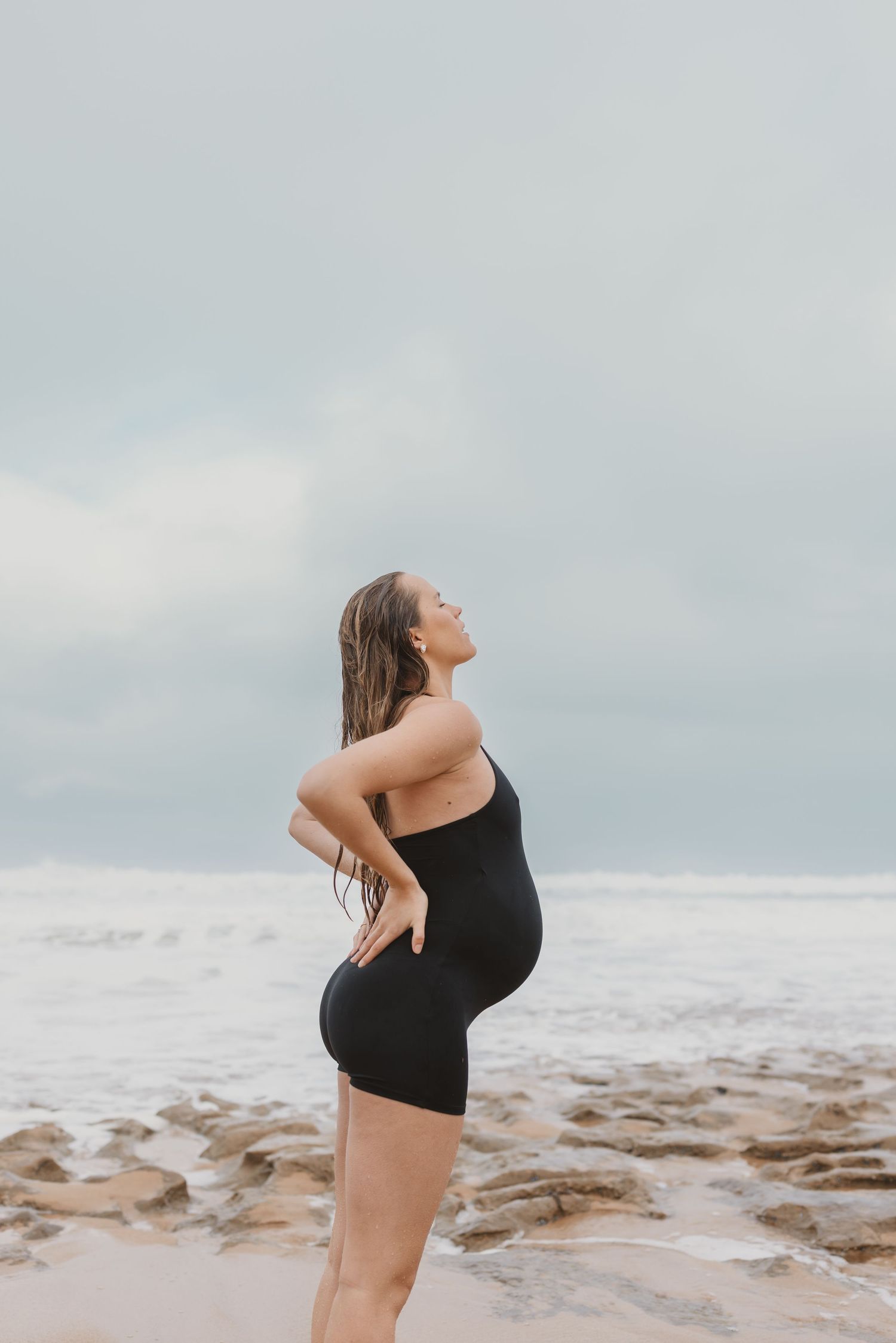 Pregnant woman in black swimsuit standing on rocky beach, hands on hips, looking up at overcast sky.