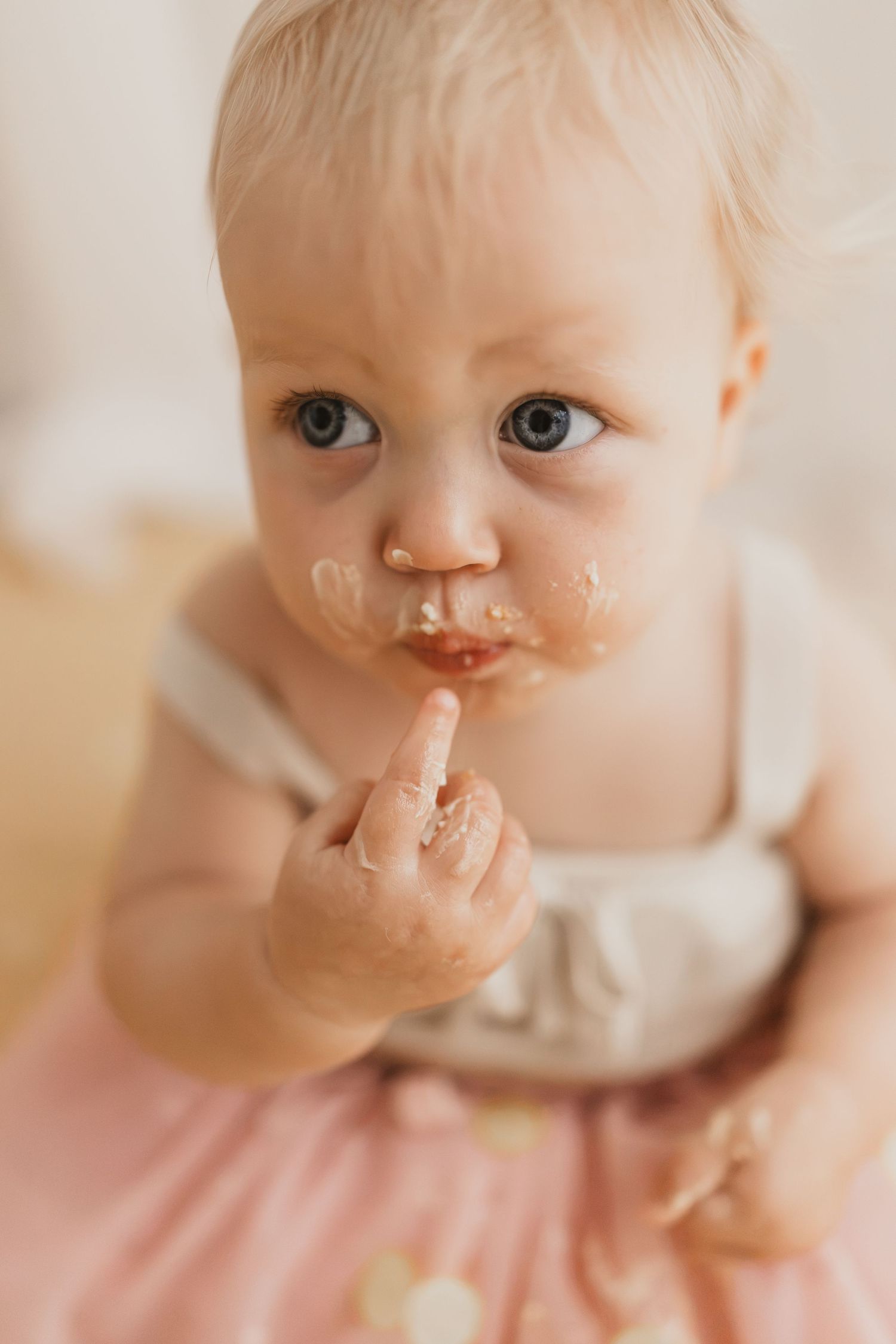 Messy baby with food on face licking finger during first birthday cake smash photo session.