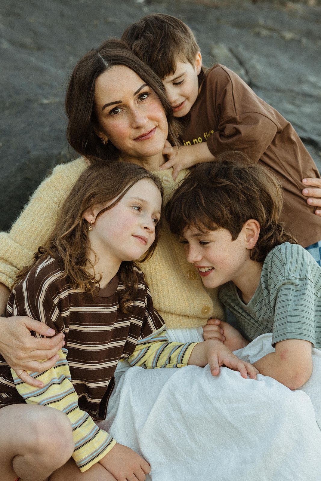 A mother in a yellow sweater sits outdoors on rocks, warmly embraced by her three young children.