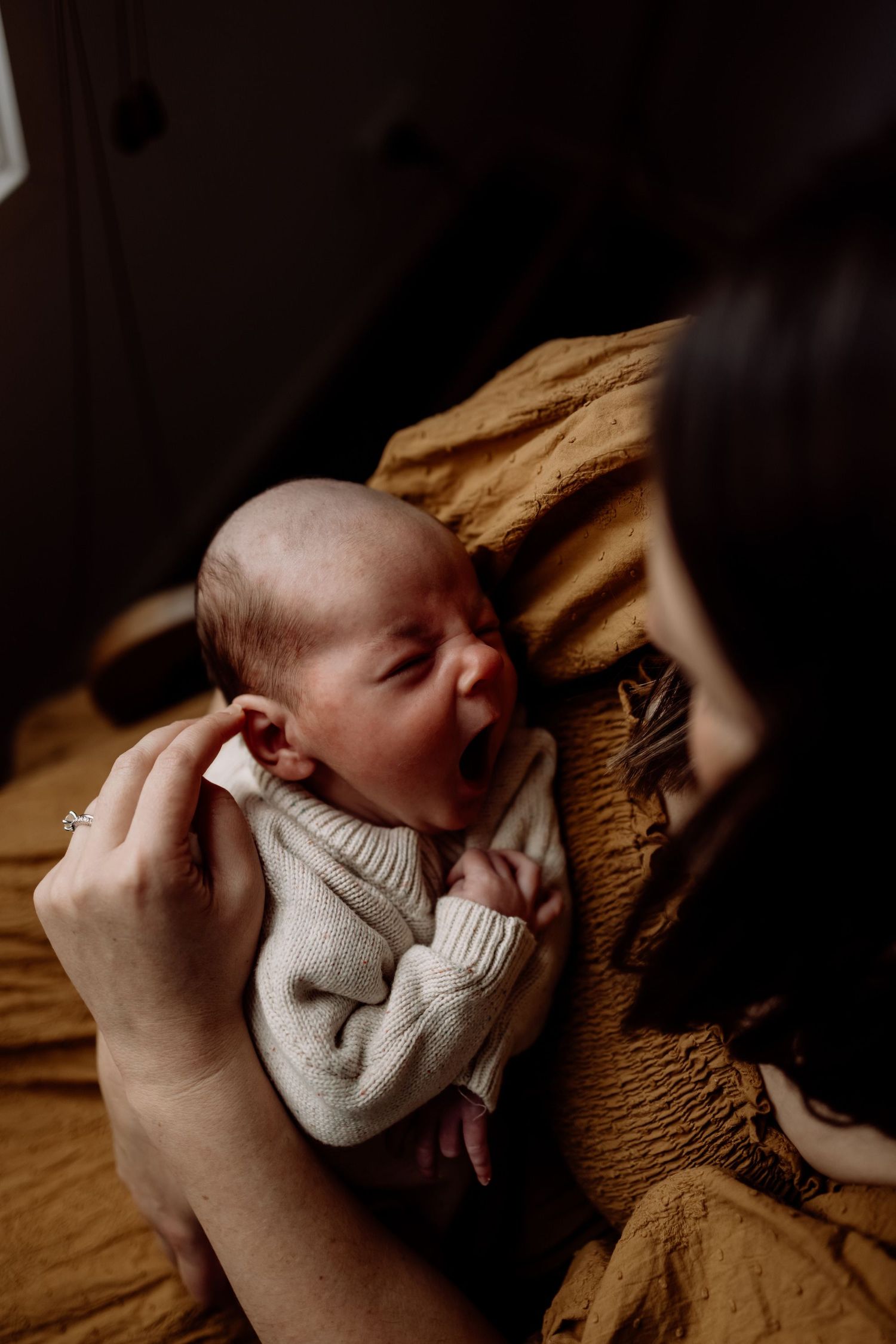 A mother tenderly holds her newborn baby in a cream knit outfit against a golden mustard blanket in warm light.