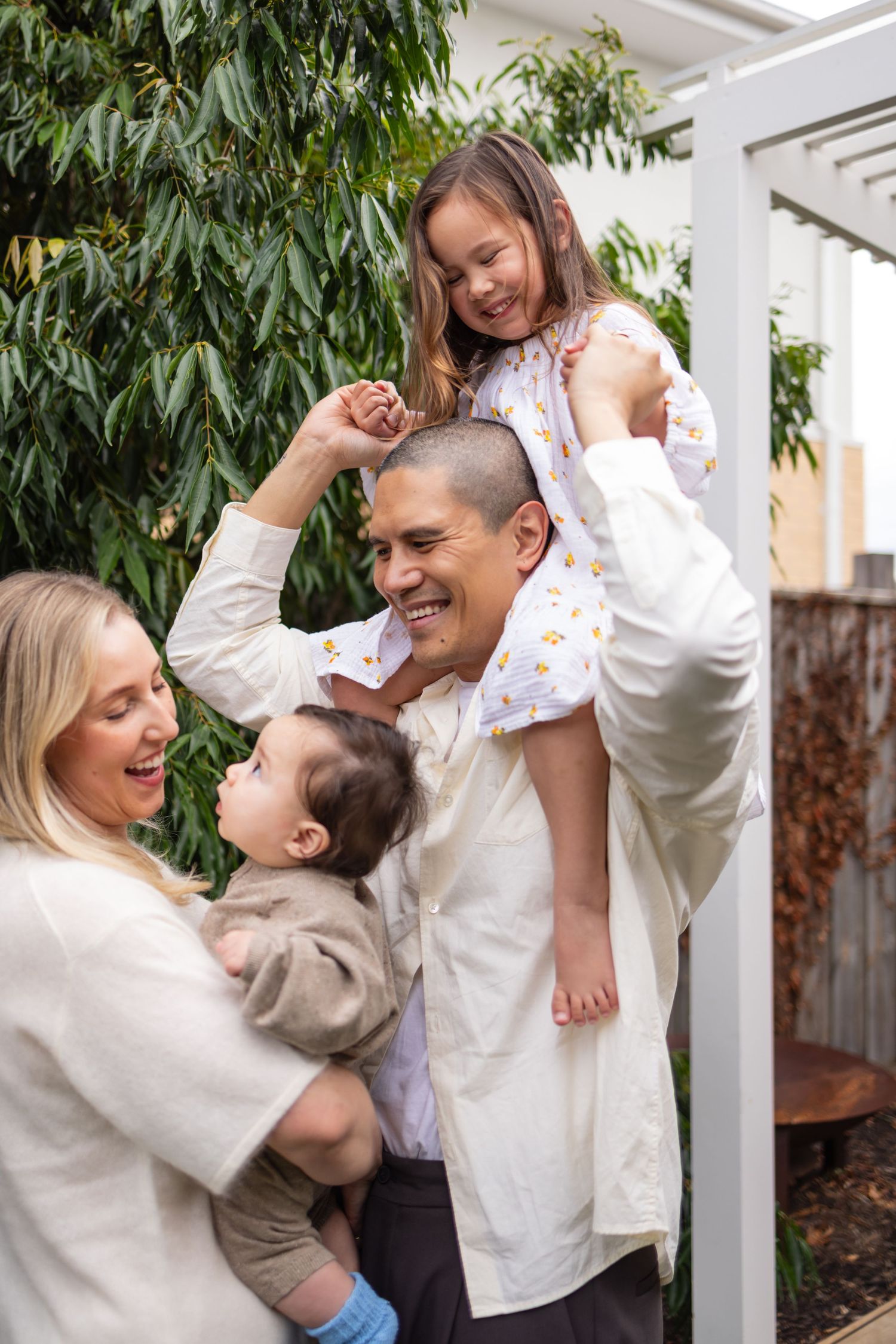 Happy family of four enjoying time outdoors, with dad carrying daughter on shoulders and mom holding baby boy.