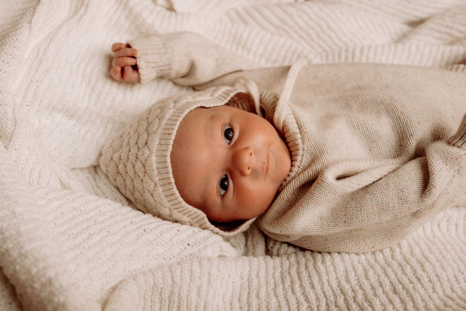 Newborn baby in cream knit sweater and bonnet lying on white blanket, peaceful and cozy.