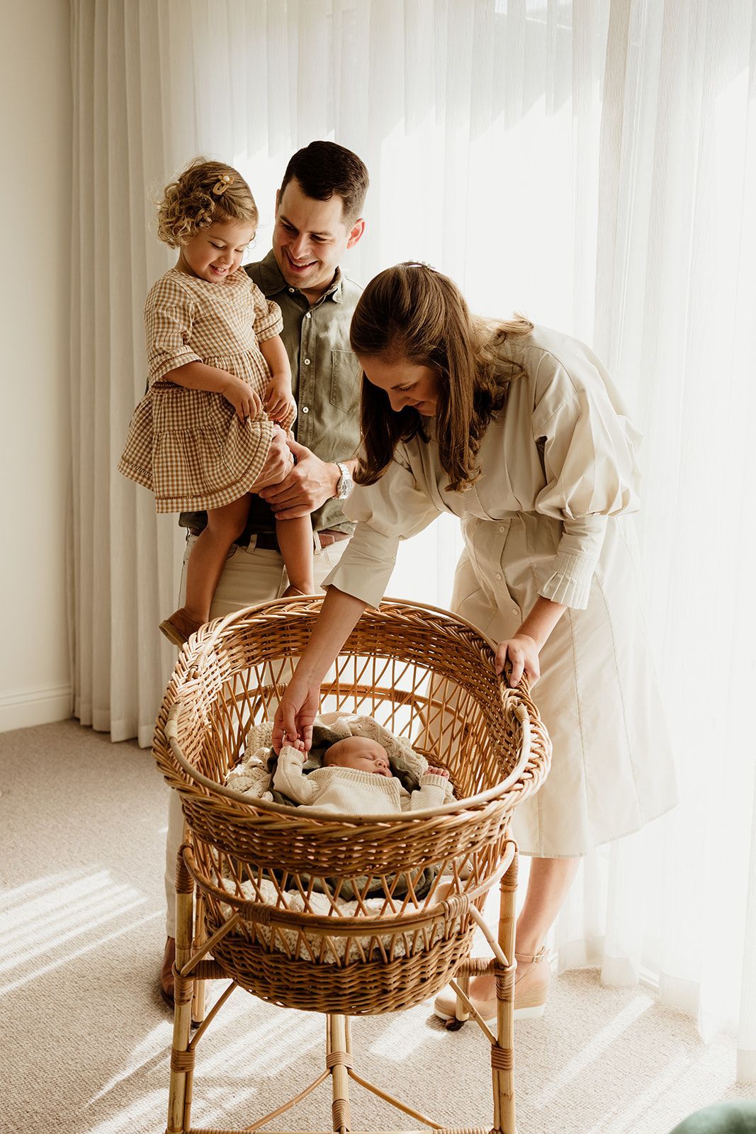 A happy family of four admires their newborn baby resting in a wicker bassinet in a bright, sunlit room.