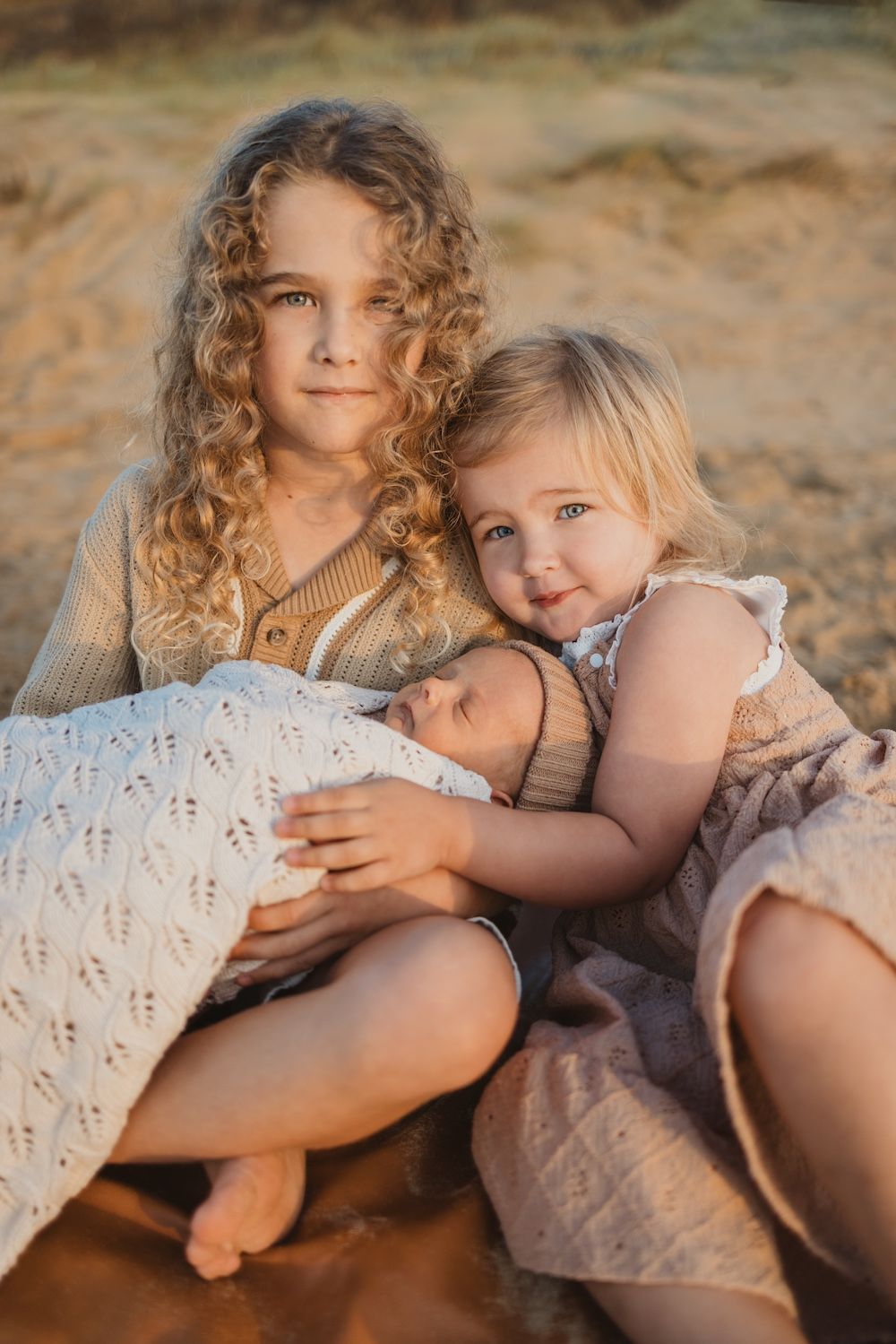 Two young girls with curly blonde hair sit on sandy beach holding a sleeping newborn wrapped in a white knit blanket.