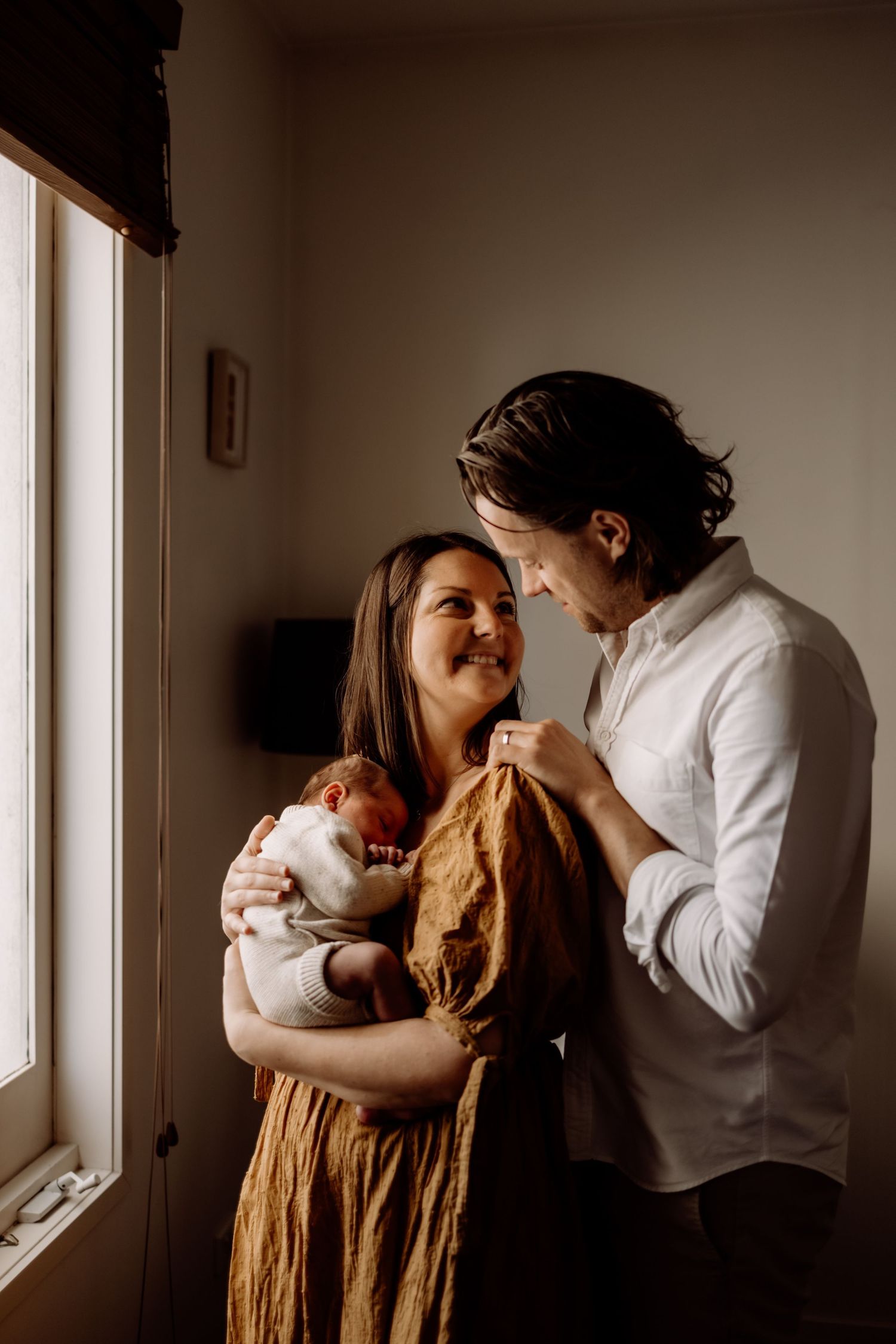 A couple smiles at each other while the mother holds their newborn baby wrapped in white near a window.