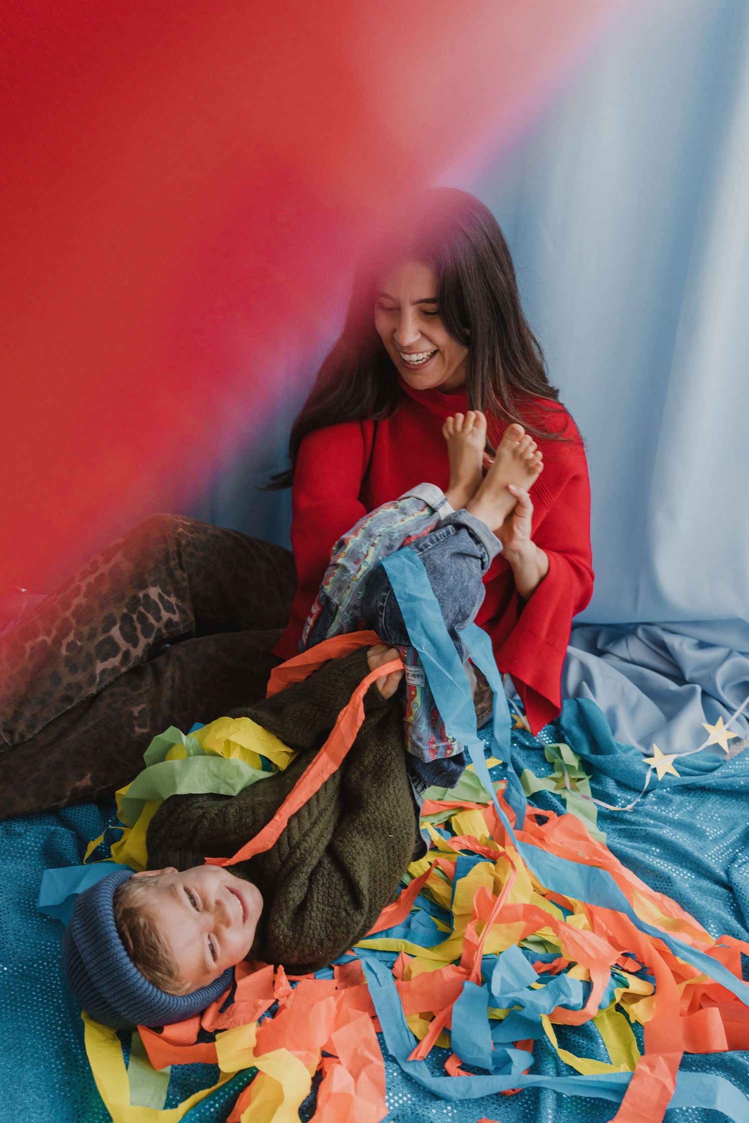 A smiling woman in a red sweater playfully holds a child's feet amid colorful ribbons on a blue blanket.