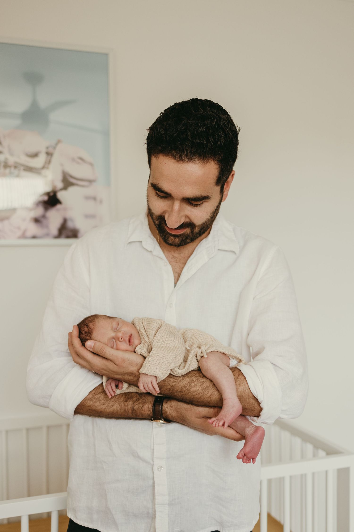 A tender moment in a nursery shows a person in white shirt holding a sleeping newborn baby against their chest.