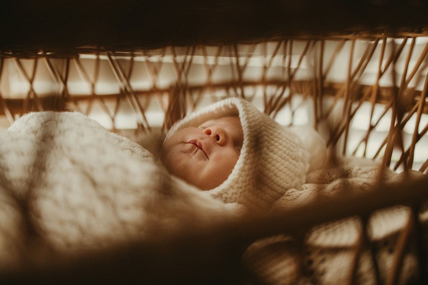 A peaceful newborn baby sleeping in a woven basket wrapped in a soft cream knit blanket and bonnet.