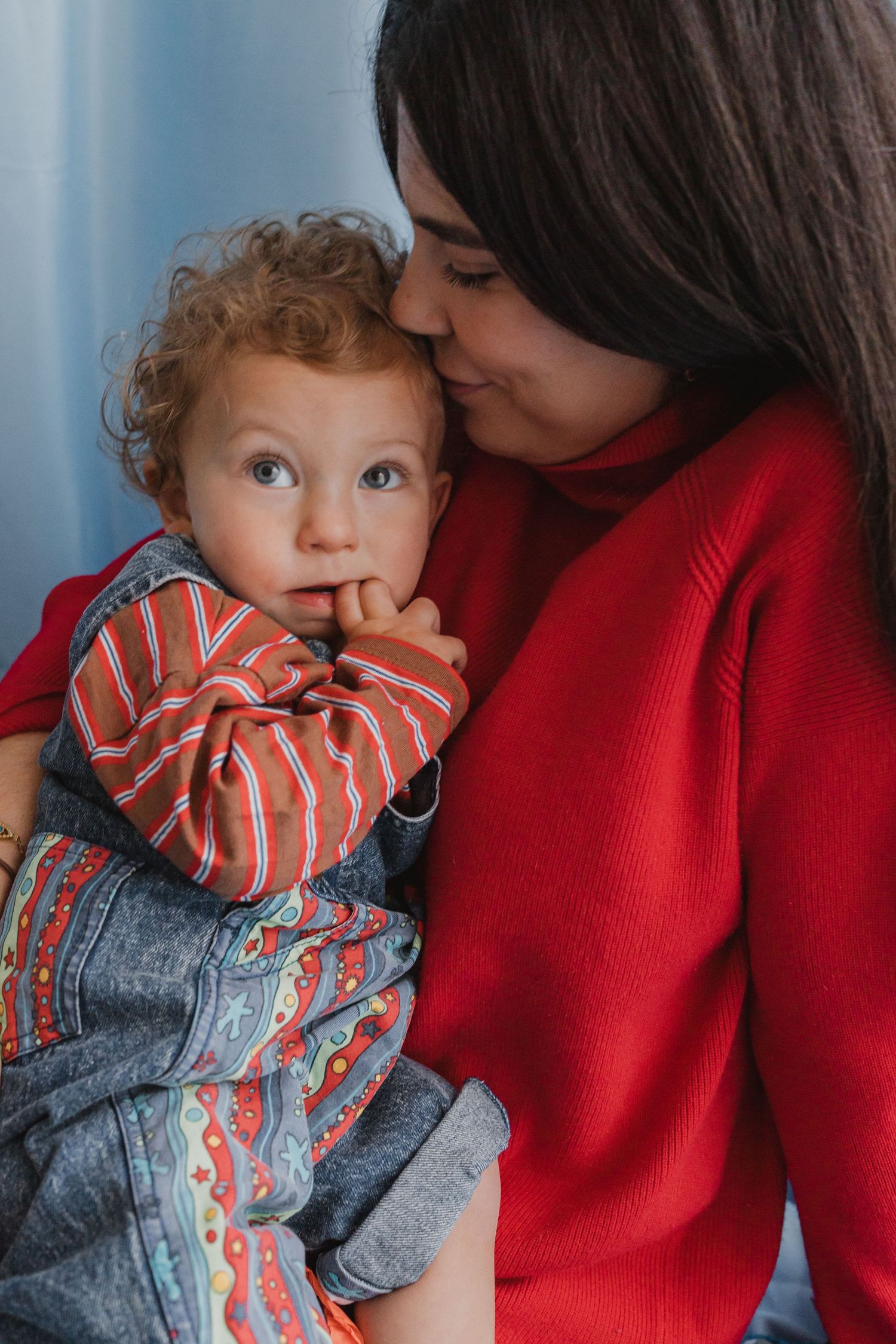 A young mother in a red sweater tenderly kisses her curly-haired toddler in a striped outfit.