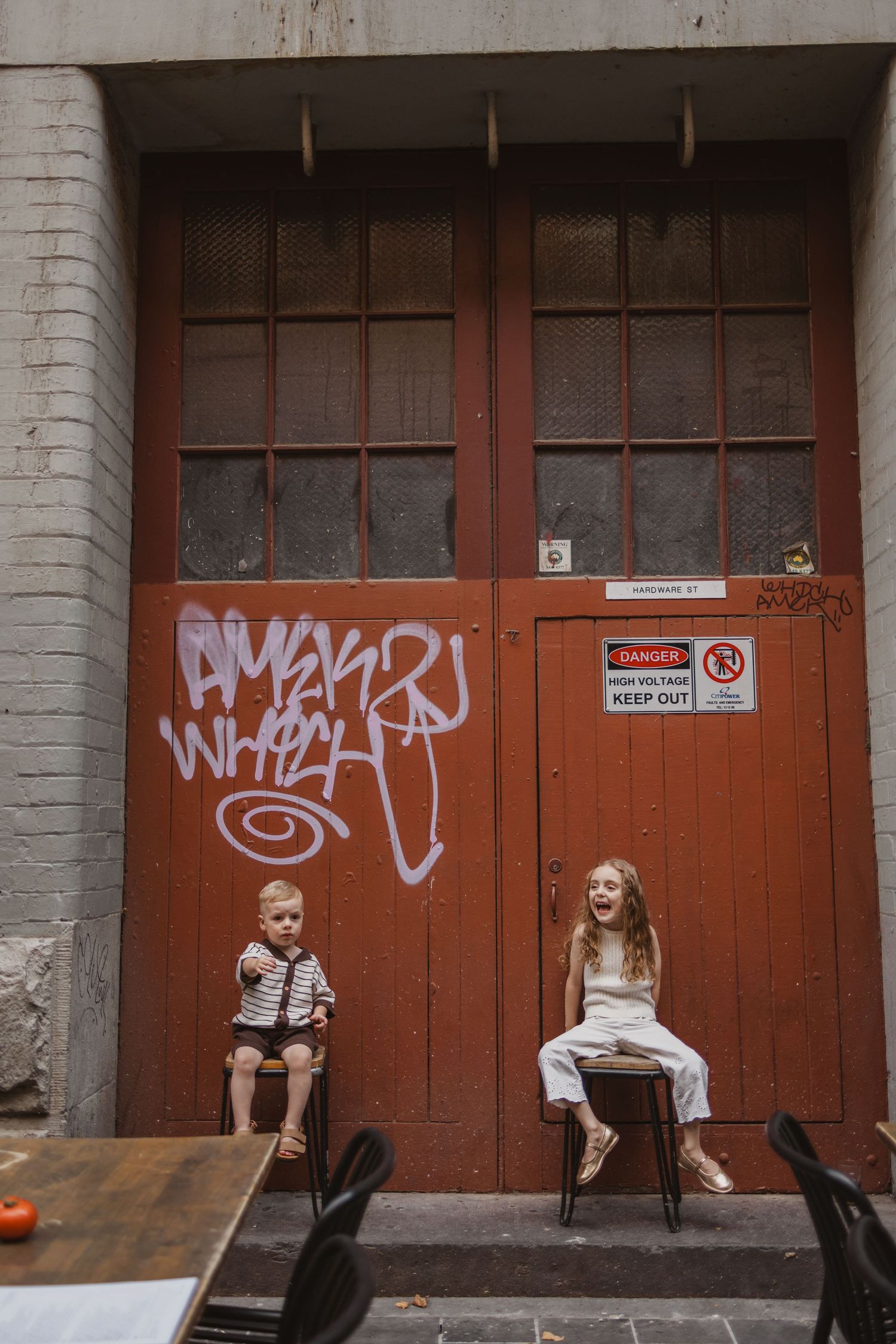 Two children sitting on chairs in front of a red graffiti-covered door in an urban alleyway.