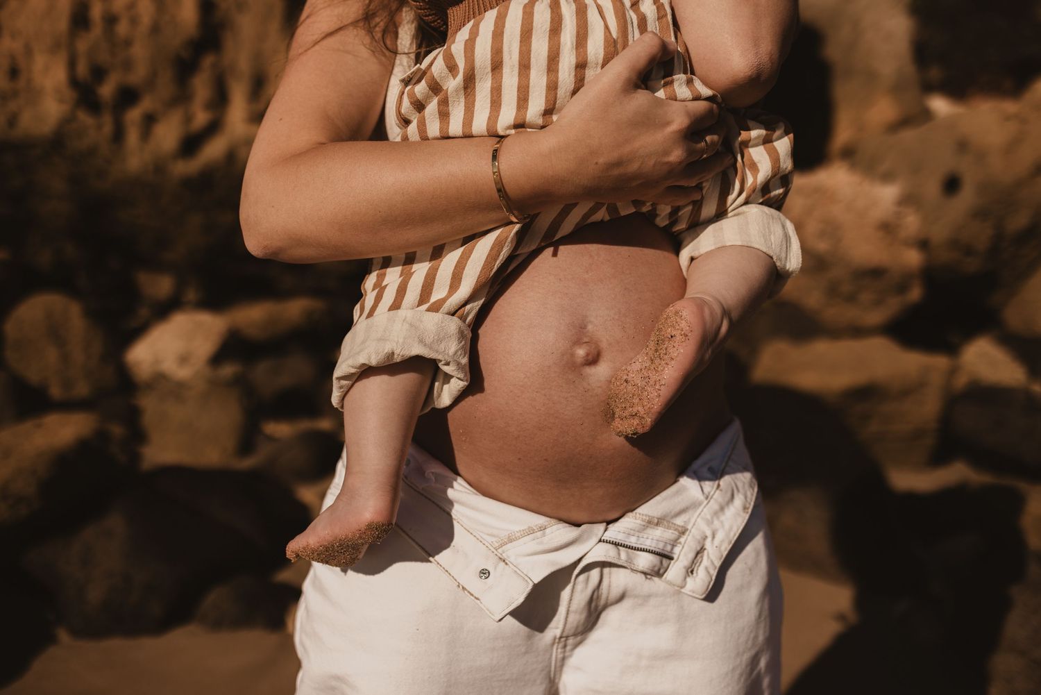 Pregnant woman in white pants holding a baby against rocky background outdoors.