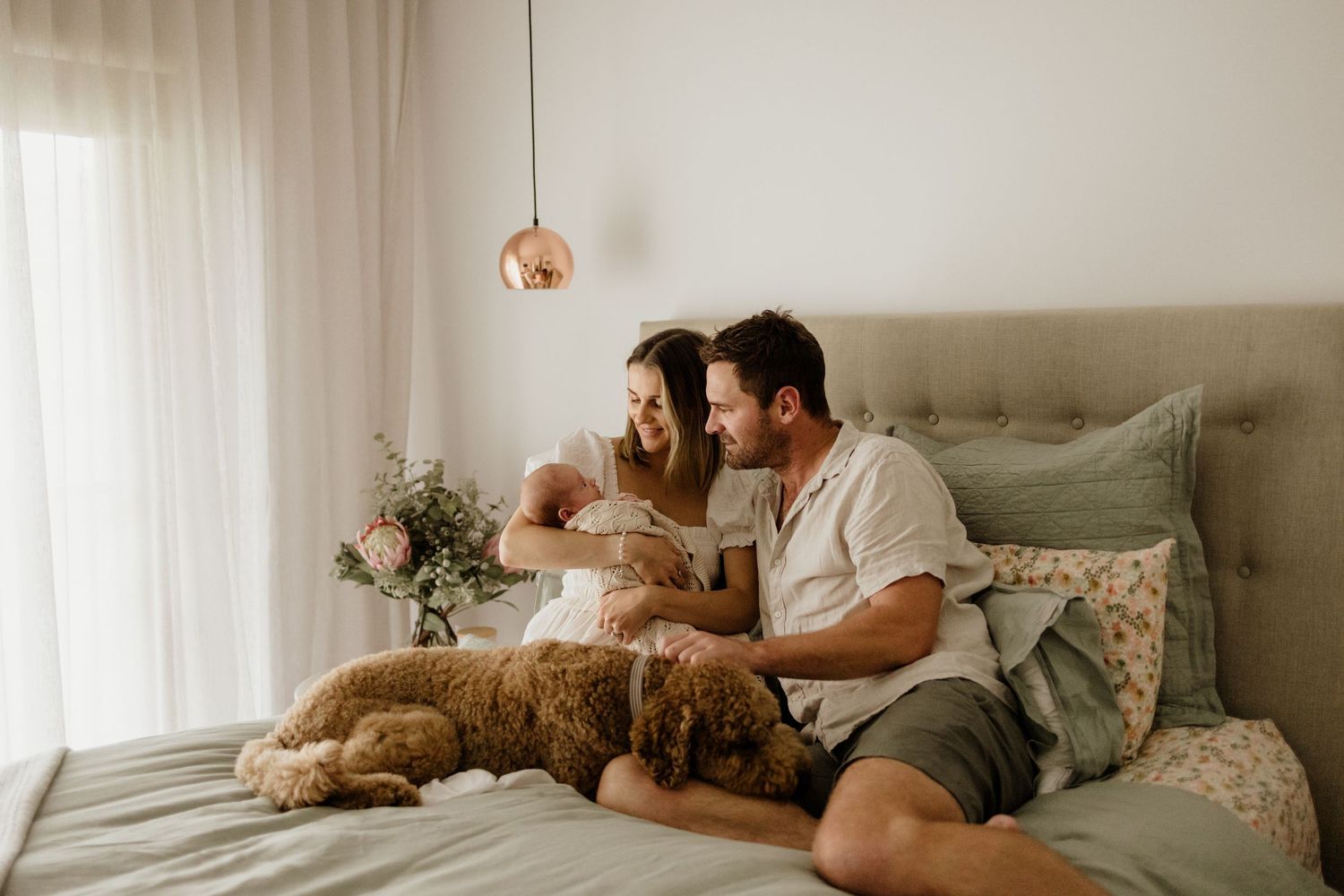 Couple sitting on a bed with a fluffy dog, holding a newborn baby in a bright, cozy bedroom.