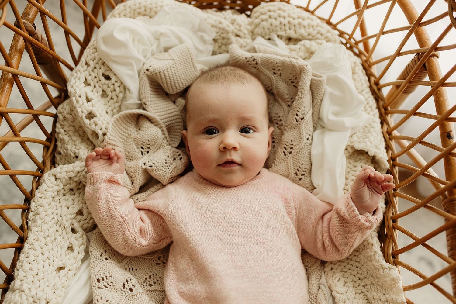 A newborn baby in a pink onesie lying in a wicker bassinet lined with a soft cream knit blanket.