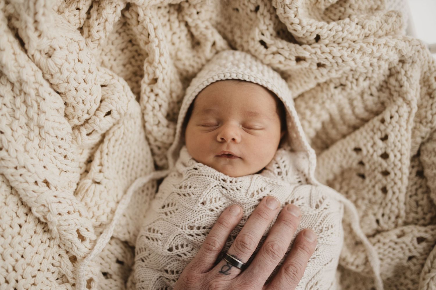 Newborn baby swaddled in cream knit blanket and bonnet, peacefully sleeping while adult hand gently holds them.