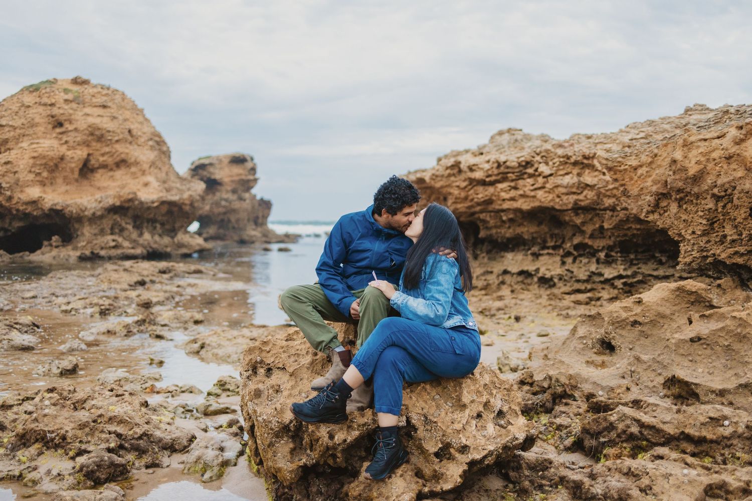A couple shares a romantic kiss while sitting on rocky coastal cliffs by the ocean on a cloudy day.