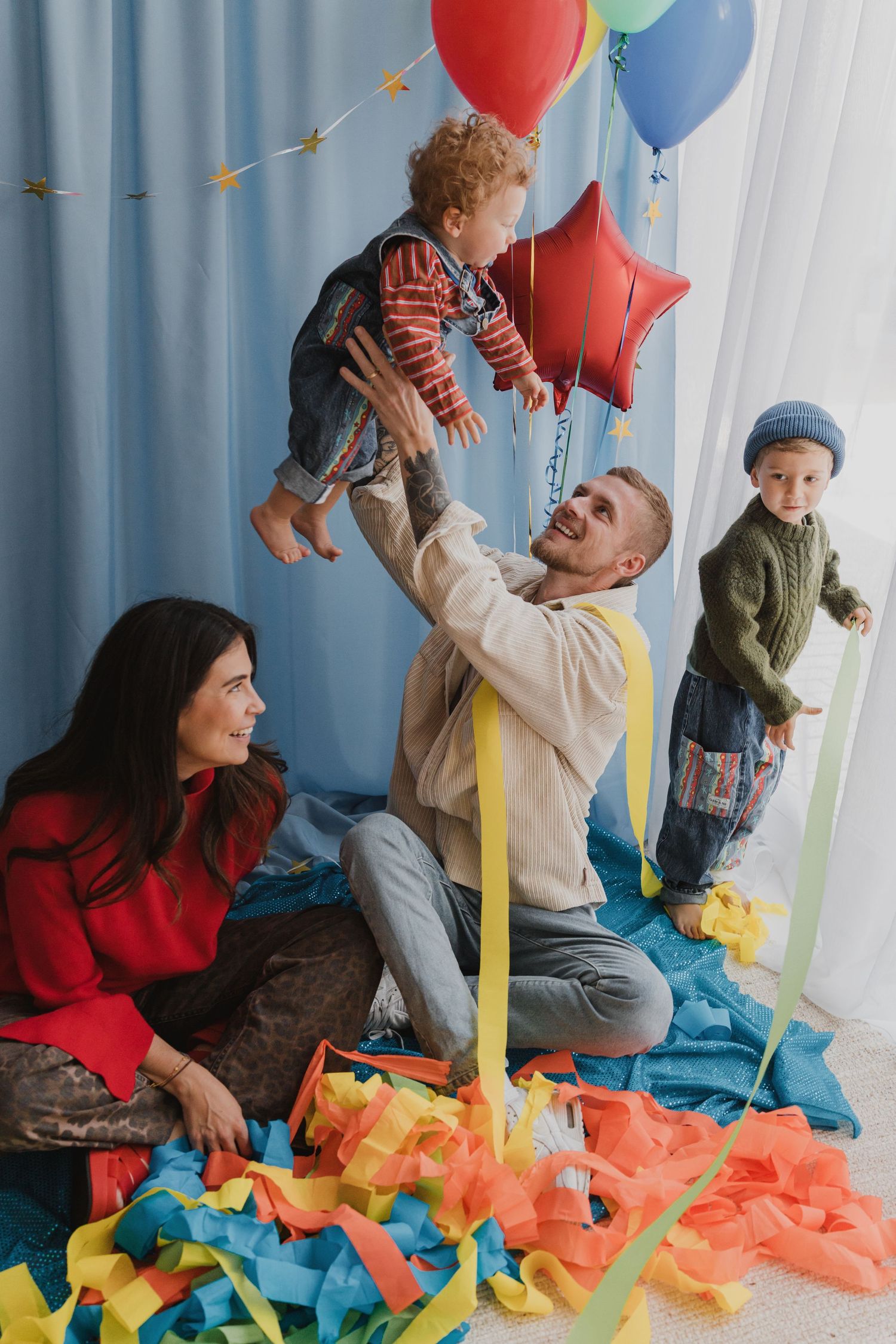 A joyful family celebrates a birthday with colorful balloons and streamers, as a father lifts his toddler in the air.