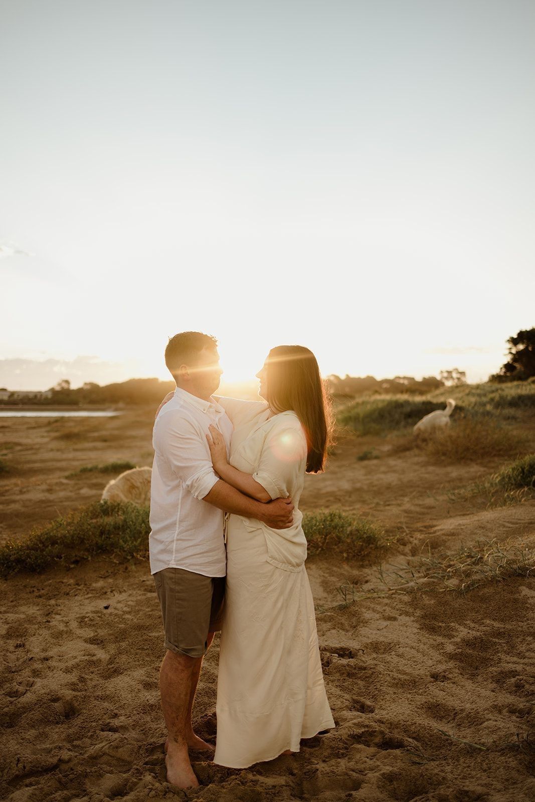 Couple embracing on sandy beach at golden sunset, woman in white dress, romantic outdoor portrait photography.