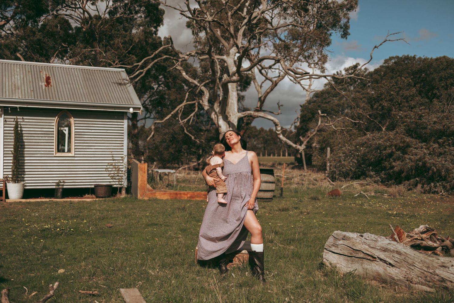 Woman in floral dress holding baby outside rustic farmhouse with gum trees in rural Australian countryside.