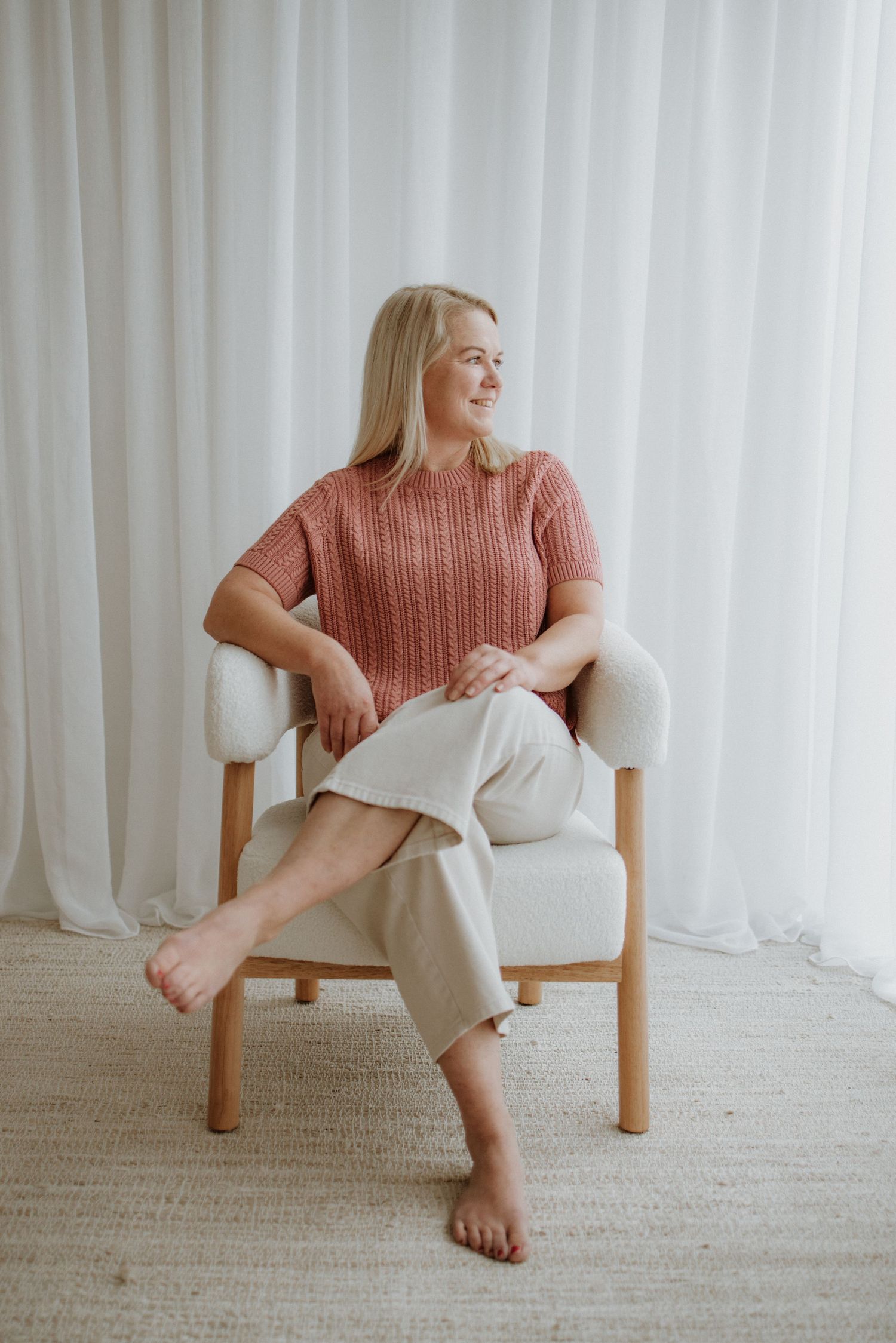 Smiling blonde woman in pink knit top and beige pants sitting relaxed in a white boucle chair by sheer curtains.