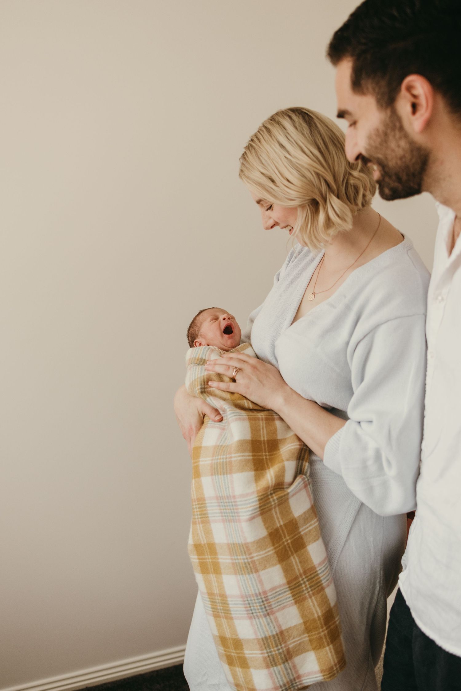 Joyful moment as parents admire their yawning newborn wrapped in plaid blanket.