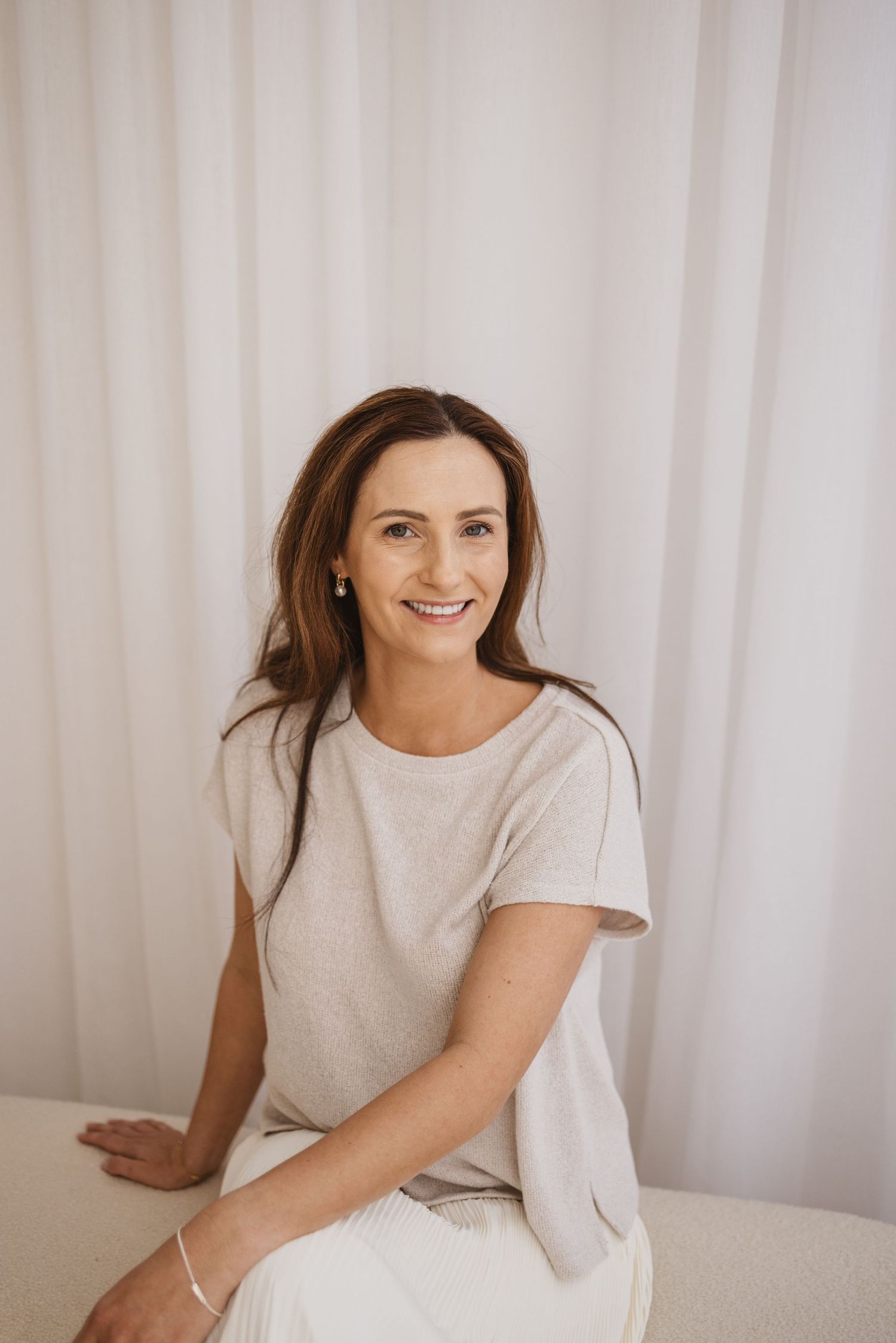 Smiling middle-aged woman with brown hair sitting in a bright white room wearing a light gray t-shirt and white pants.