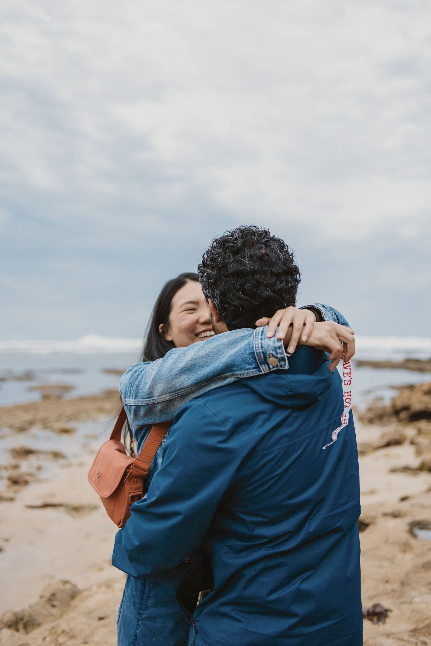 A smiling woman hugs a man in a blue jacket on a rocky beach under a cloudy sky.