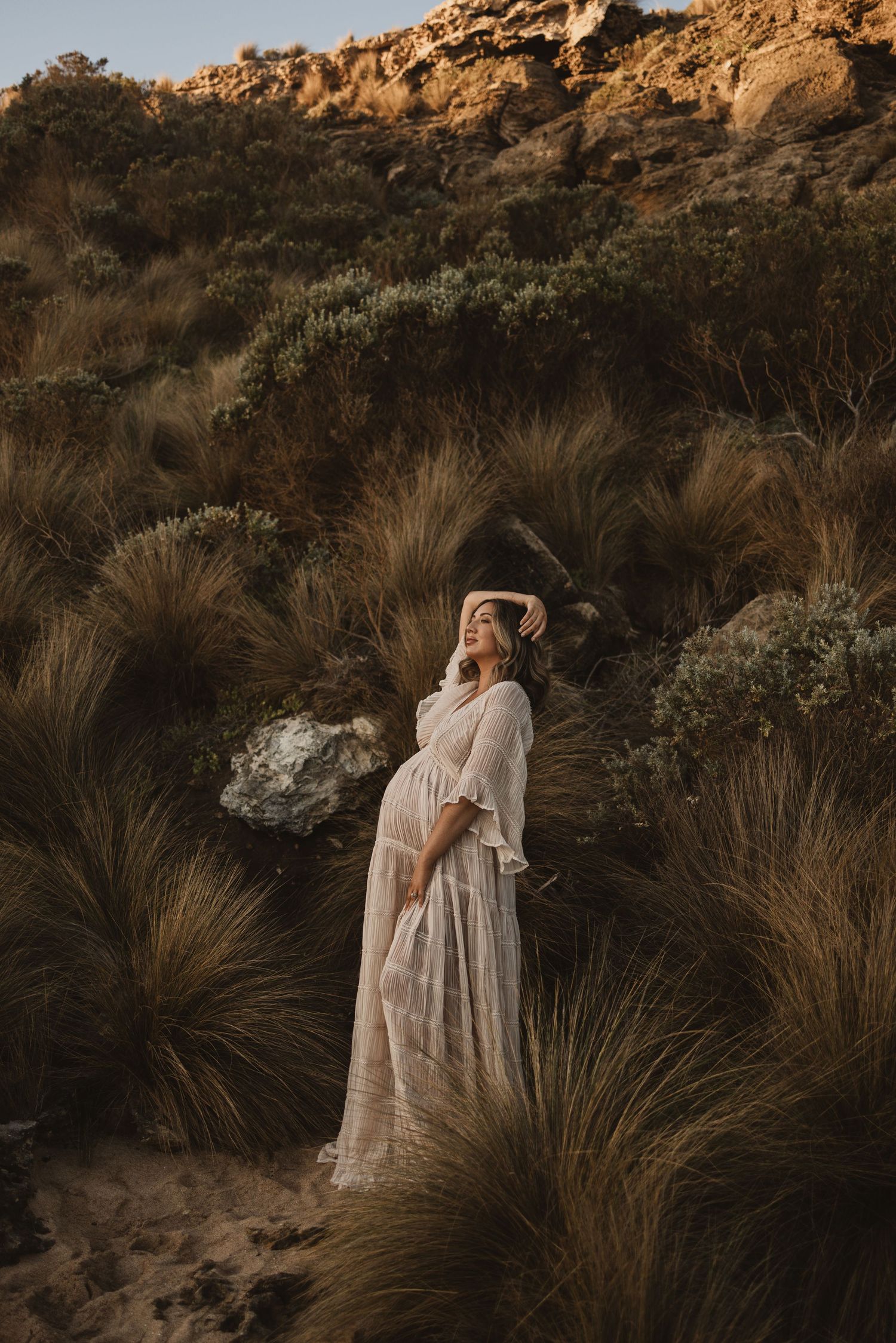Pregnant woman in white lace dress standing among tall grasses and rocky hillside at golden hour.
