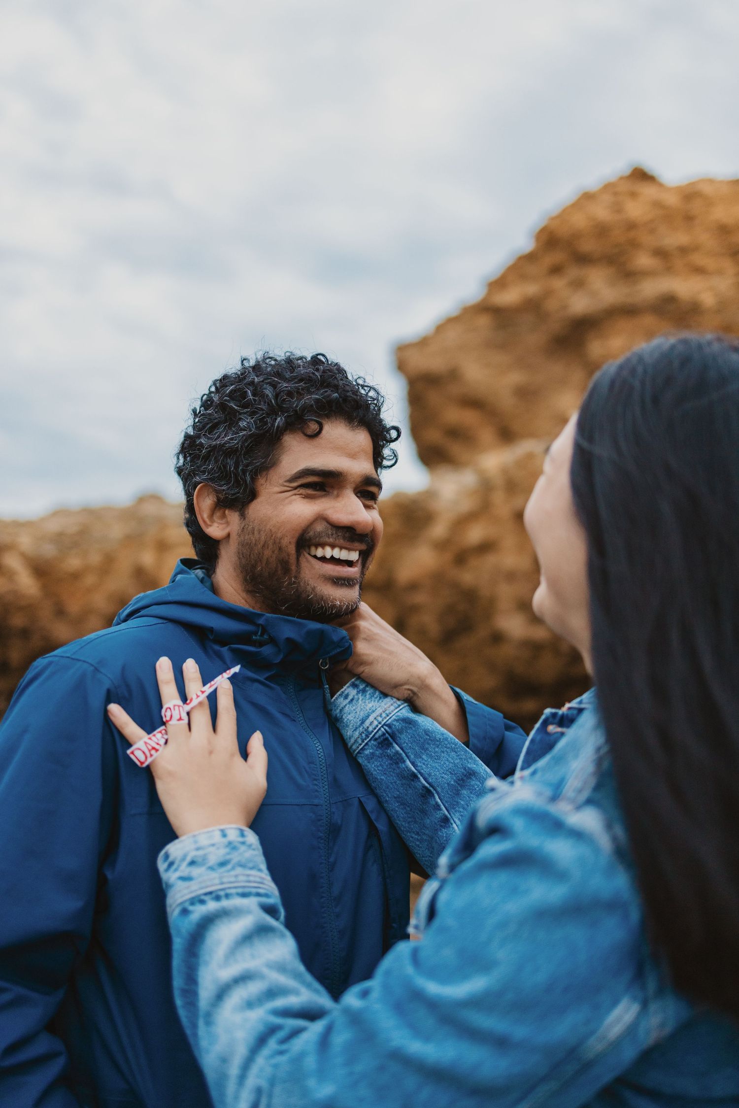 A smiling man in a blue hoodie interacts with a woman in a denim jacket outdoors near rocky cliffs.