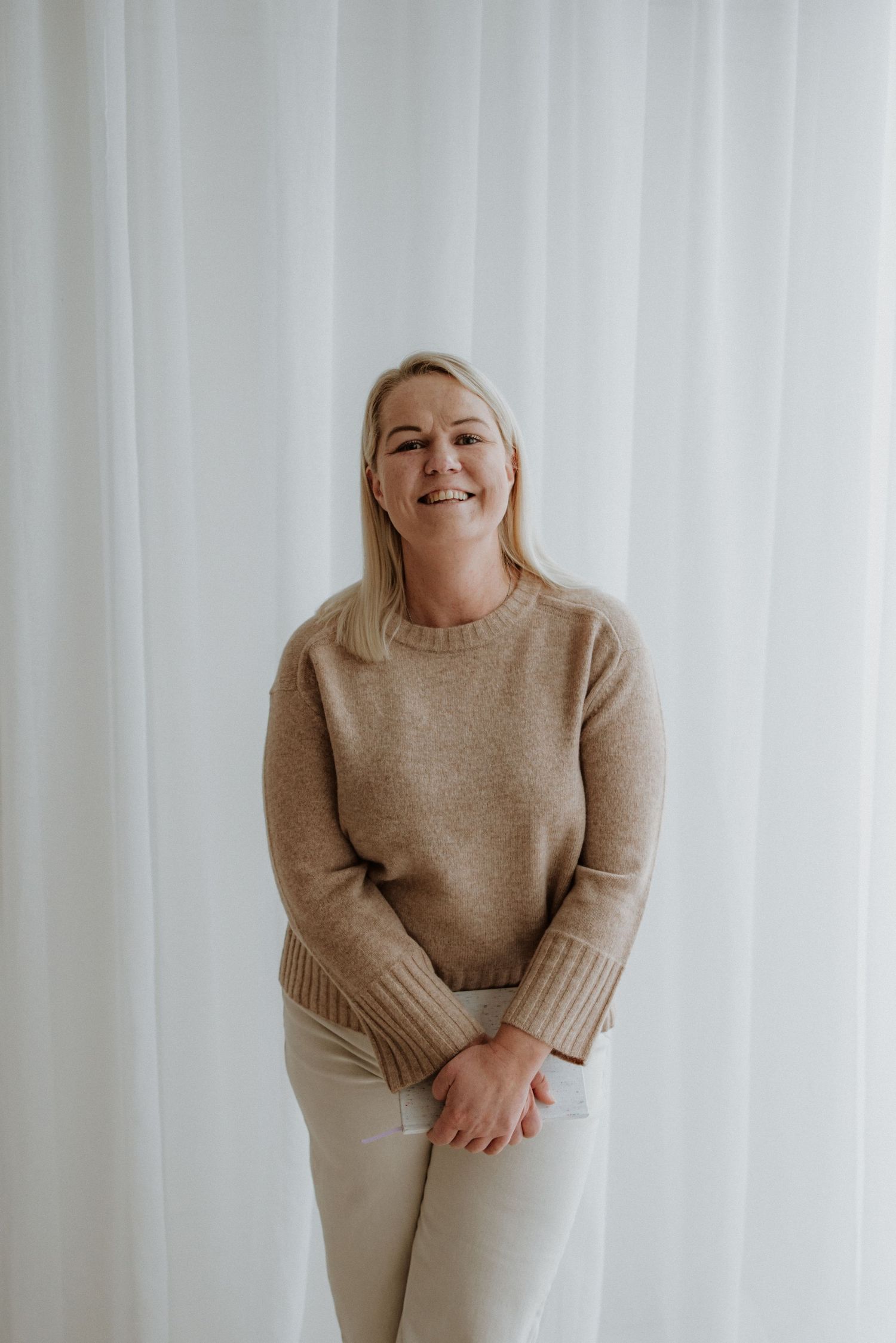Smiling blonde woman in beige knit sweater and white pants standing against white curtain backdrop.