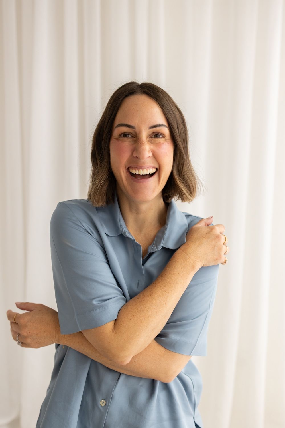 Smiling woman in blue polo shirt hugging herself, laughing joyfully against white curtain background.