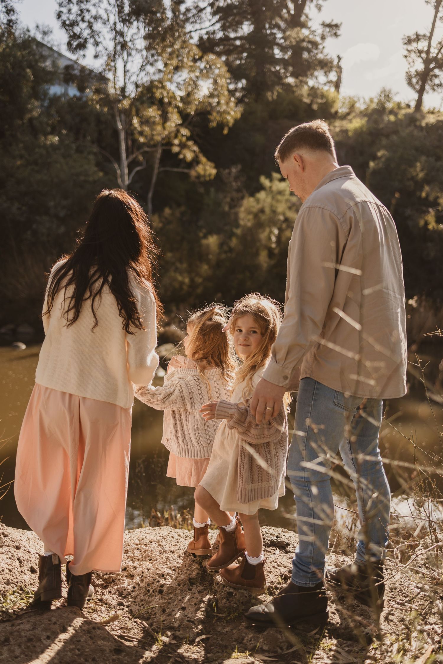 Family of four standing outdoors near a river, parents and two young girls in matching cream dresses, golden hour light.