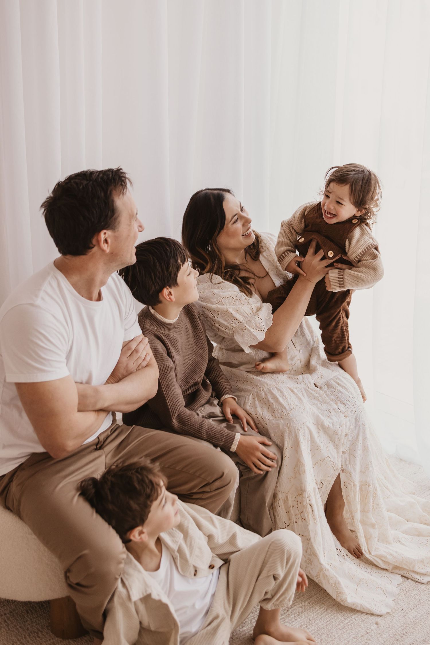 A happy family of five poses together in a bright, airy room with sheer curtains in neutral, earthy tones.