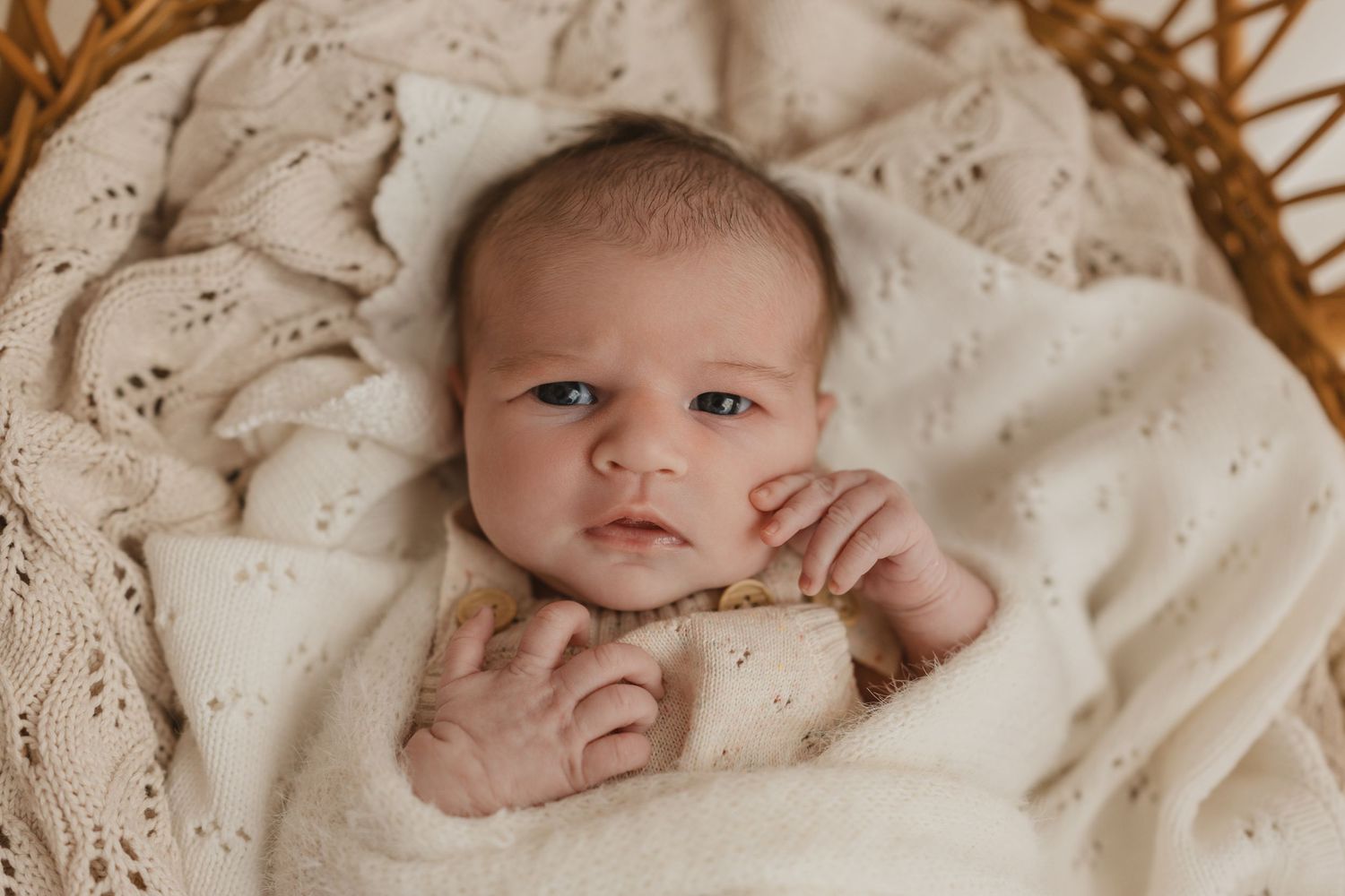 A newborn baby with blue eyes lies in a wicker basket wrapped in a soft cream knit blanket.