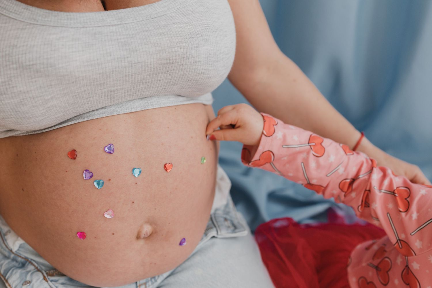Pregnant woman with colorful heart gem stickers decorating her bare baby bump, wearing a white crop top and jeans.